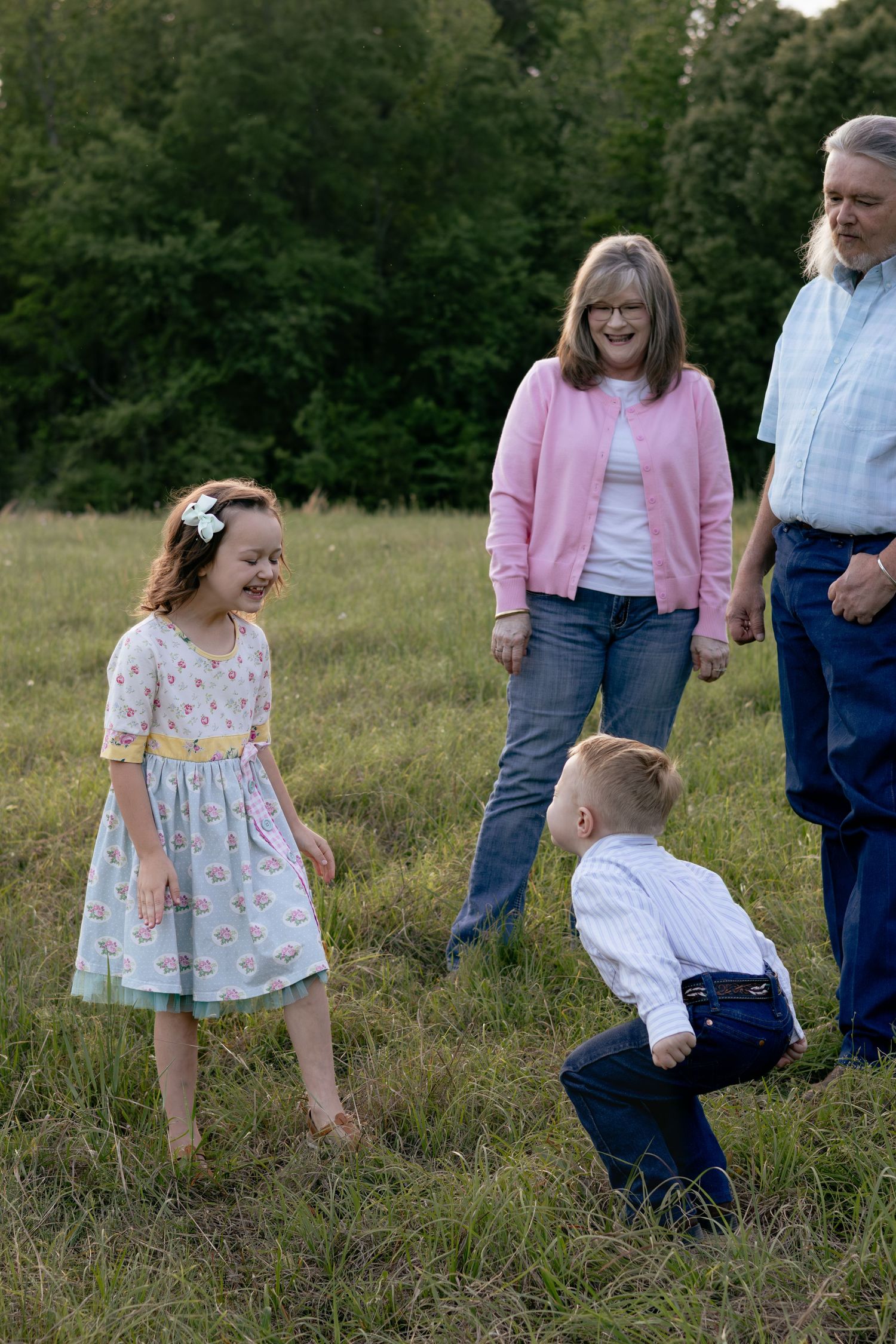 Grandparents watching two young children play in a green meadow, girl in floral dress laughing at toddler boy.