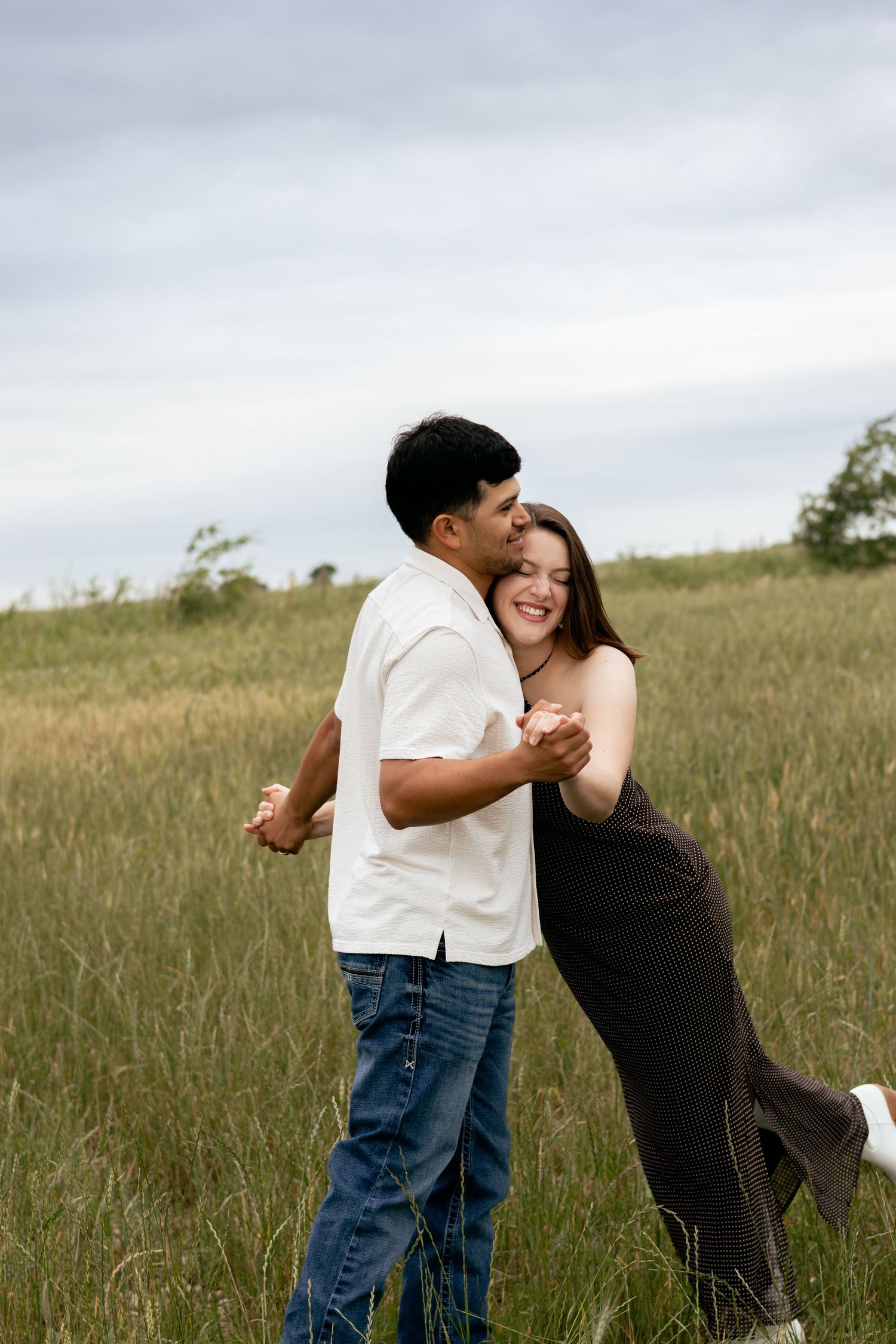 Happy couple dancing and laughing together in a grassy meadow, woman in black dress leaning back joyfully.