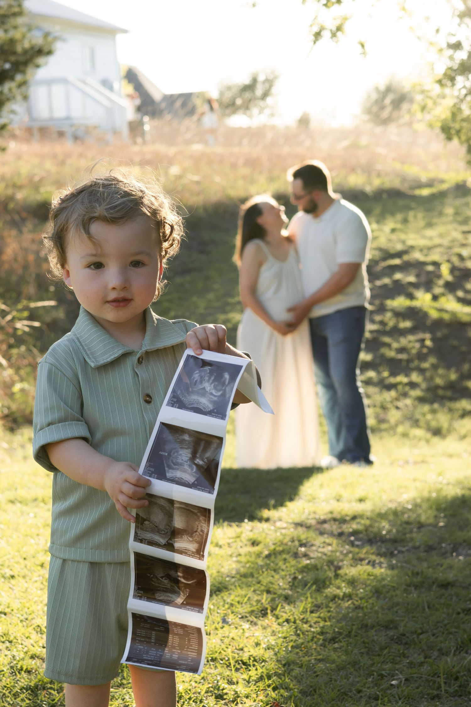 A toddler holds ultrasound photos outdoors while expecting parents embrace in the background during a pregnancy announcement shoot.