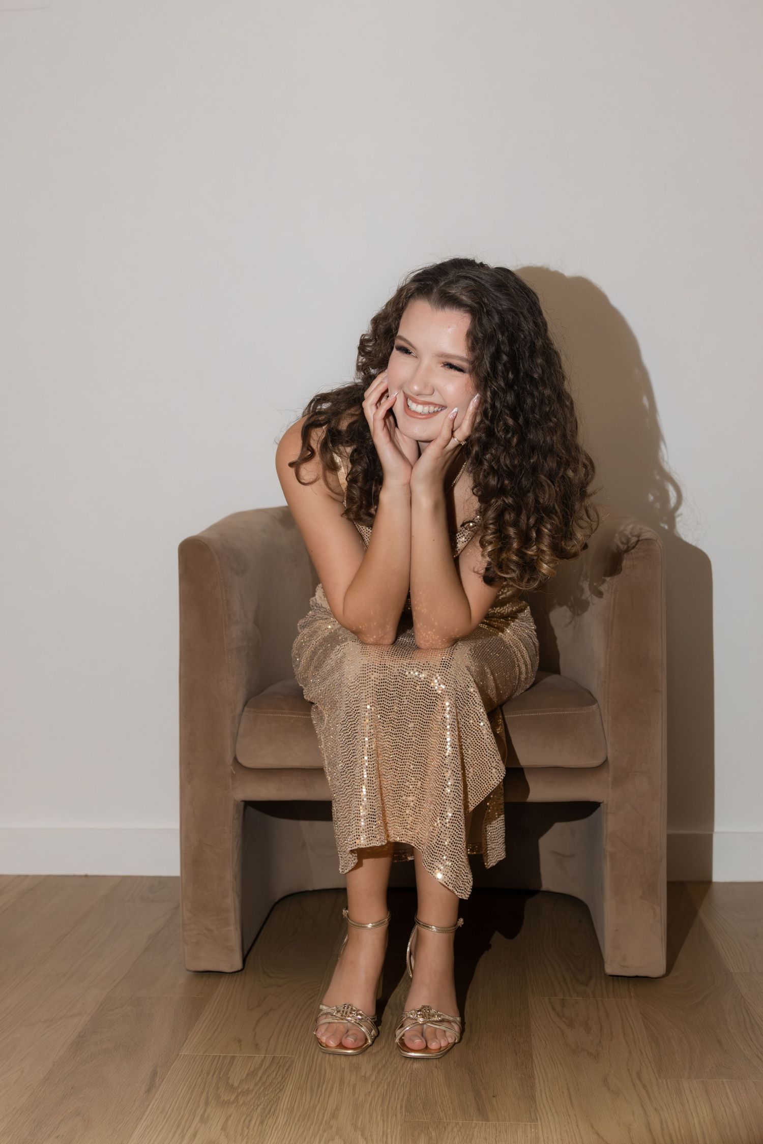 Smiling young woman in gold sequin dress sitting in beige velvet chair with hands on cheeks.