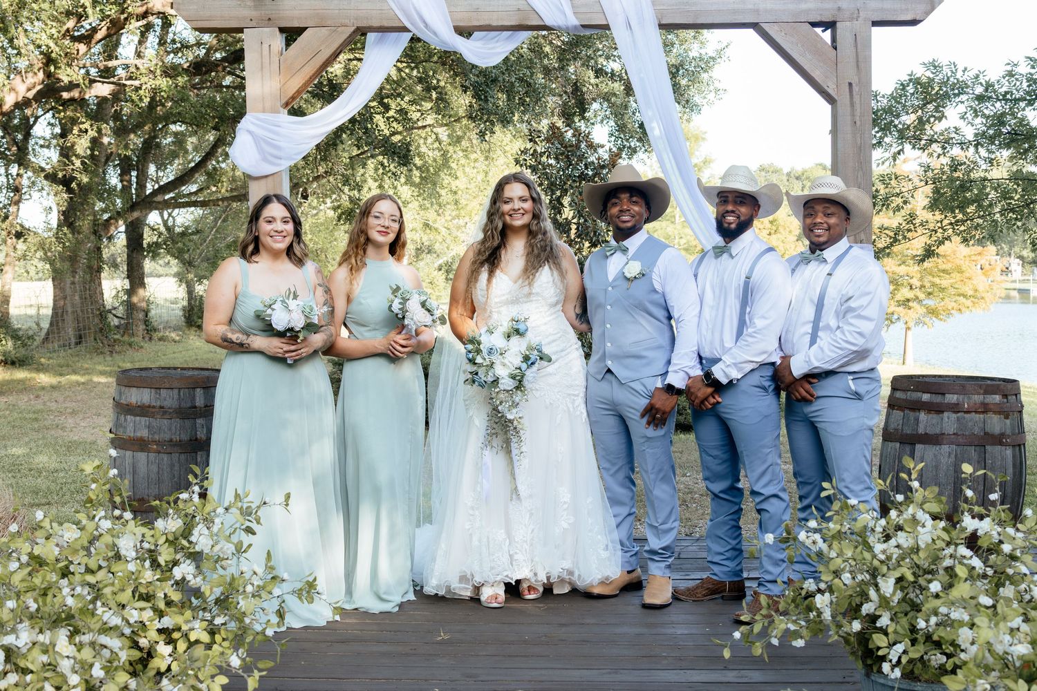 Bride in white gown with bridesmaids in sage green dresses and groomsmen in blue suits at outdoor rustic wedding ceremony.