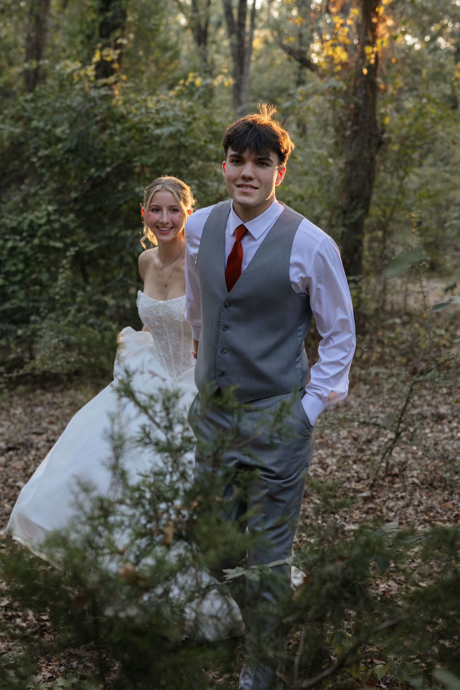 Smiling groom in gray vest and red tie leads bride in white gown through lush green woodland setting.