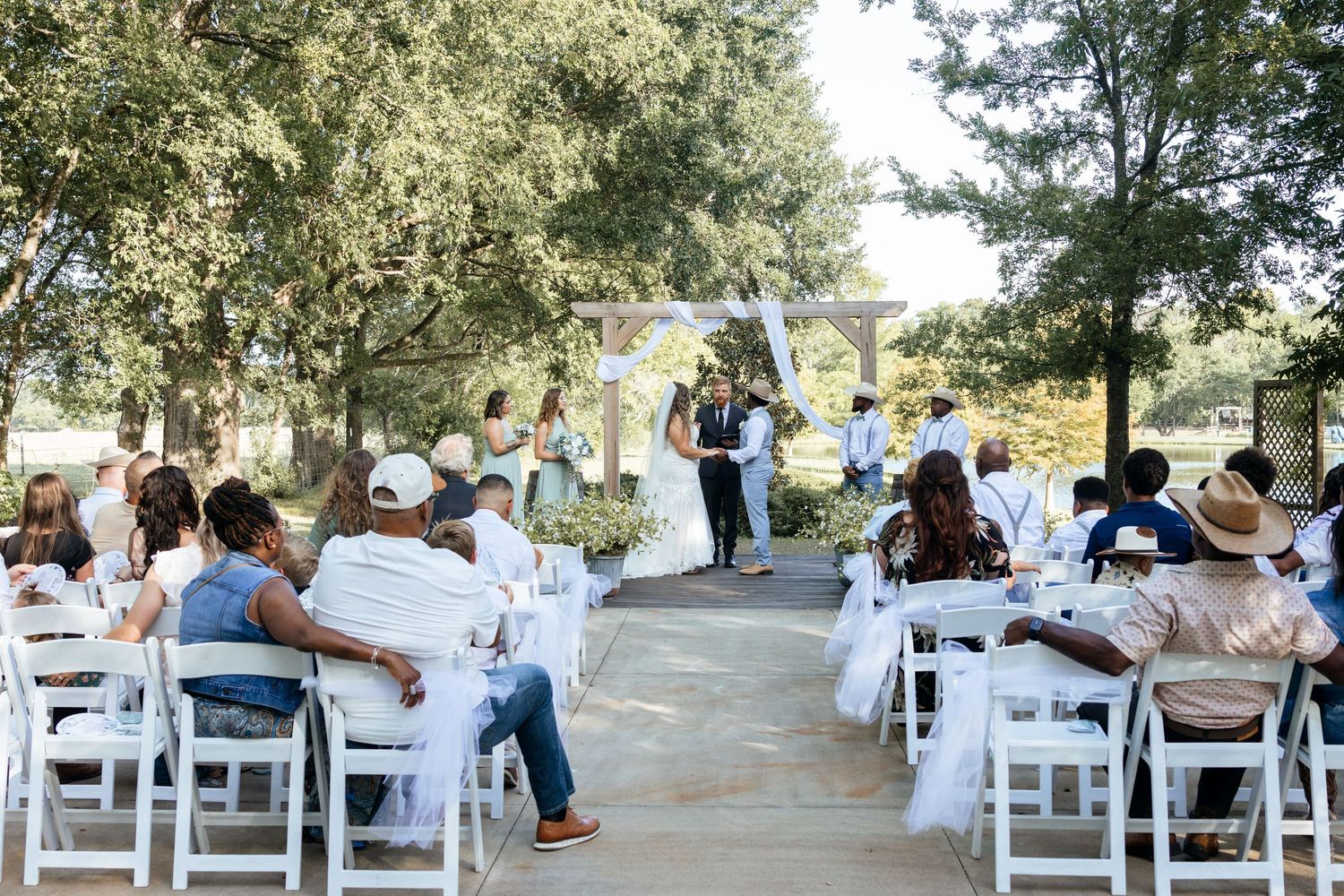 Outdoor wedding ceremony with guests seated in white chairs under trees, couple exchanging vows at floral arch altar.