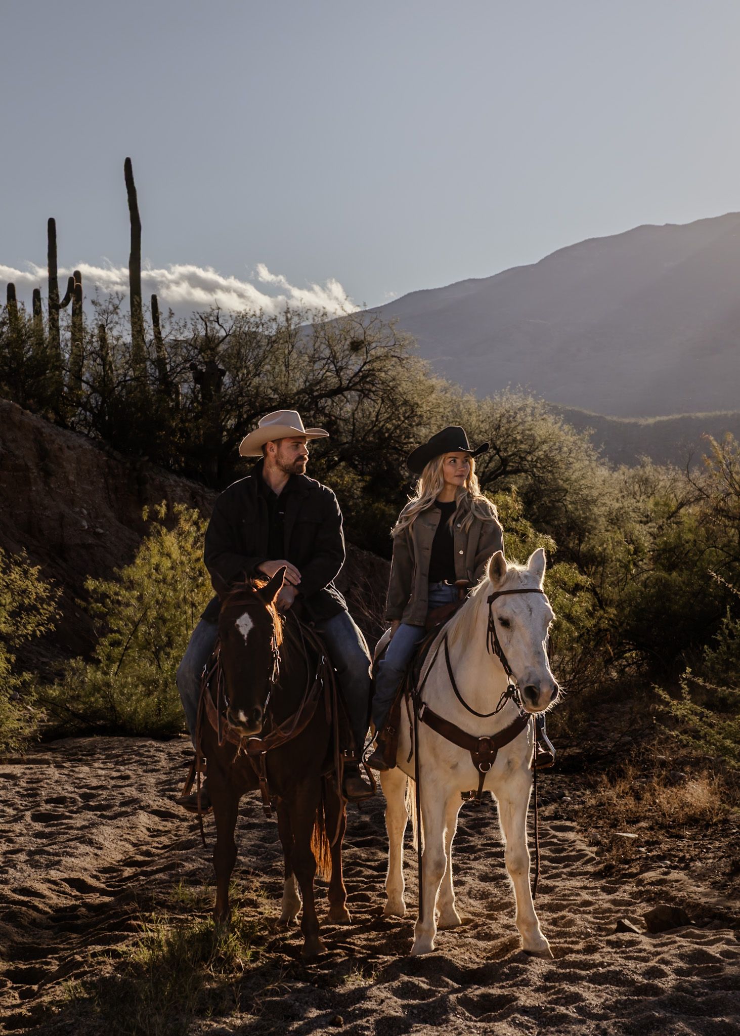 Horses and riders traverse rocky desert terrain with mountains and saguaro cacti in the background.