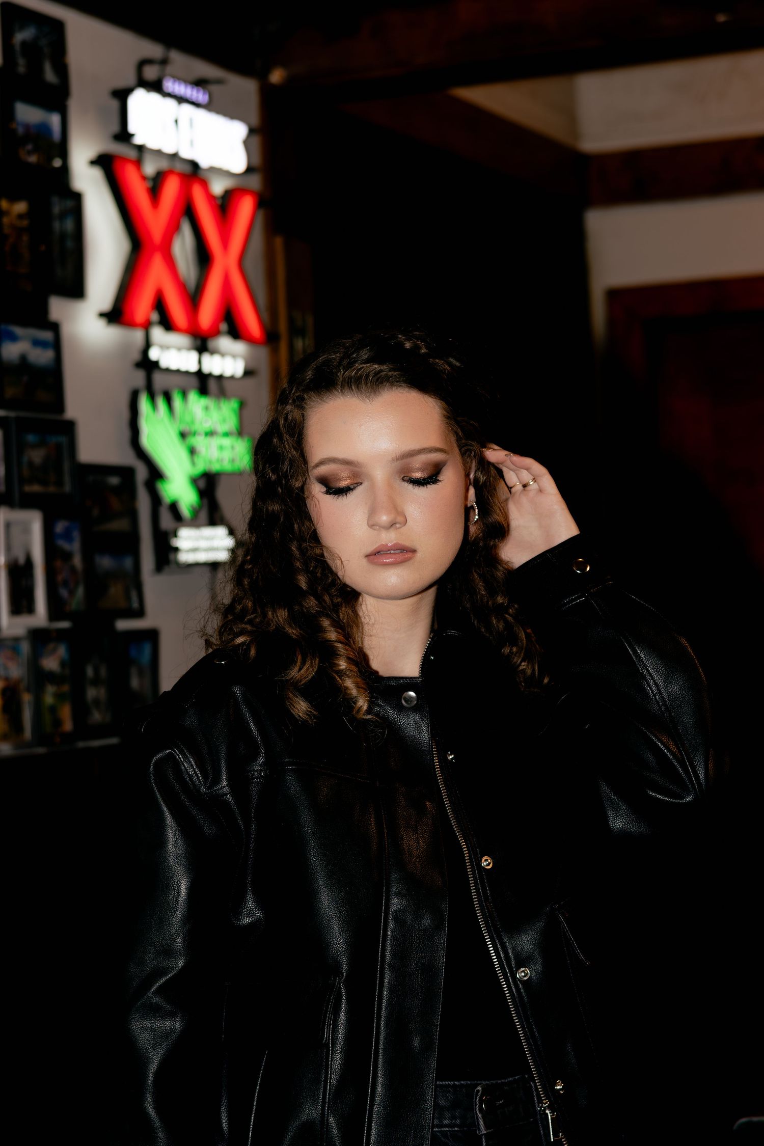 Young woman in black leather jacket poses under neon XX sign in dimly lit bar setting.