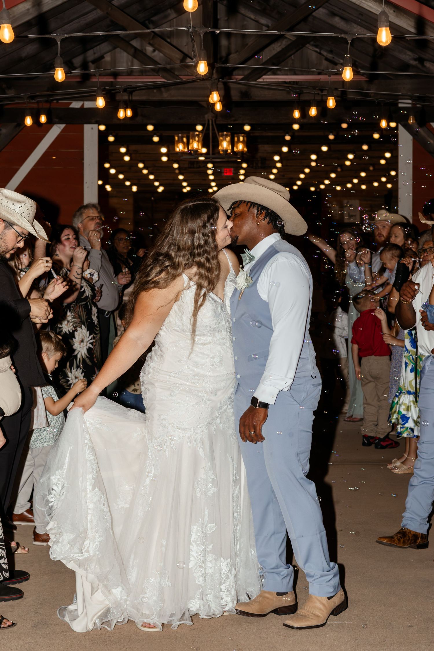 A bride and groom share a kiss at their rustic barn wedding reception, surrounded by cheering guests and warm string lights.
