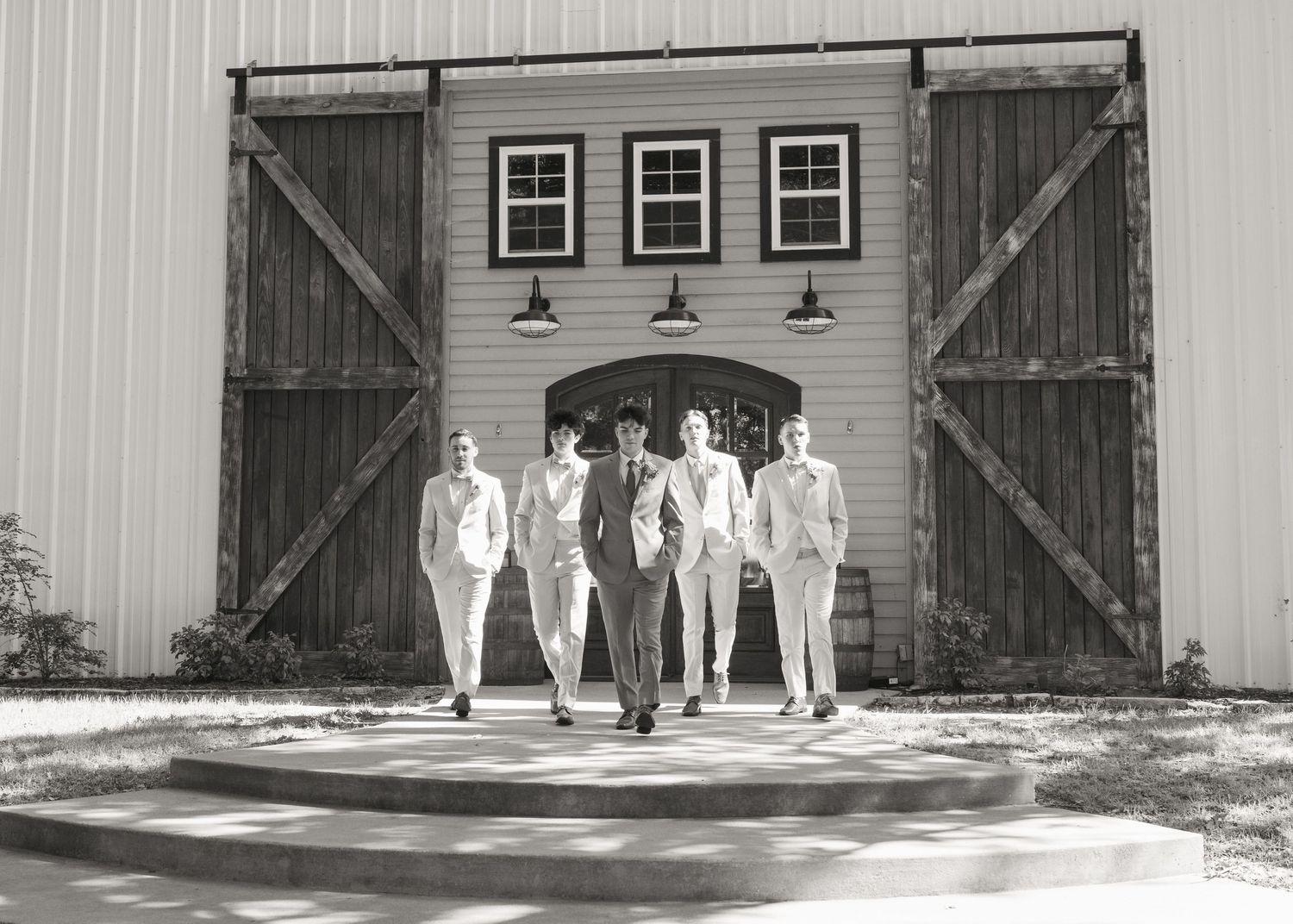 Five groomsmen walking confidently in front of rustic barn doors in black and white wedding photo.