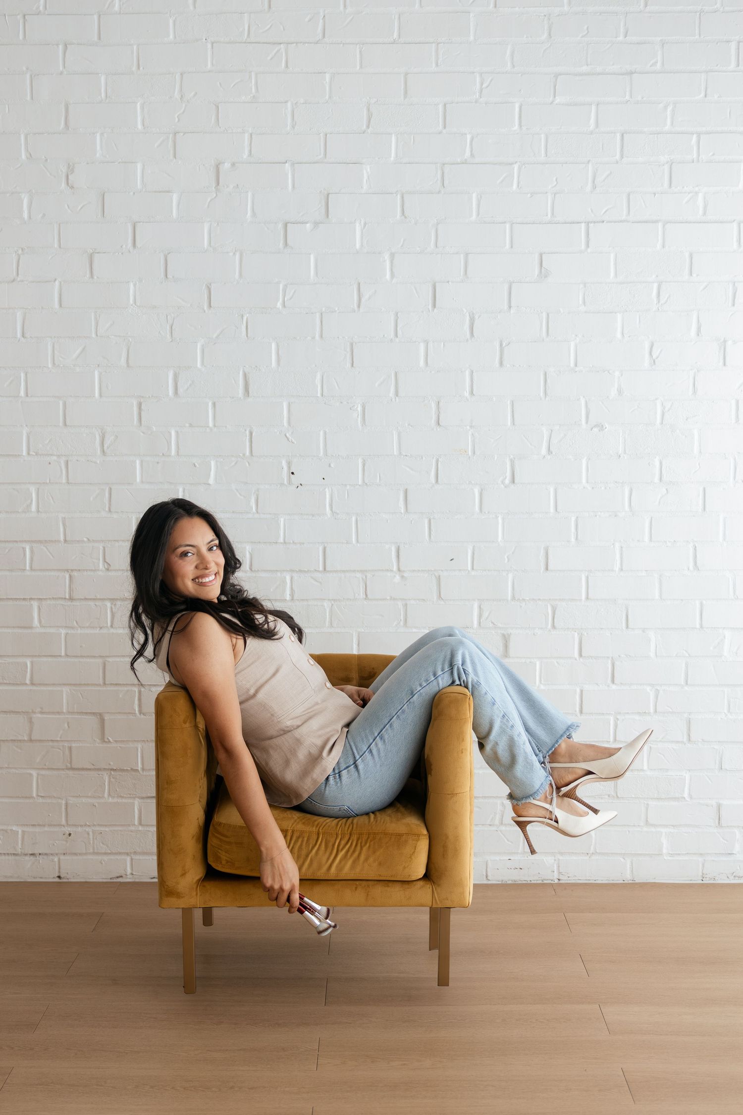 Woman in beige top and jeans relaxing in mustard yellow armchair against white brick wall, smiling.