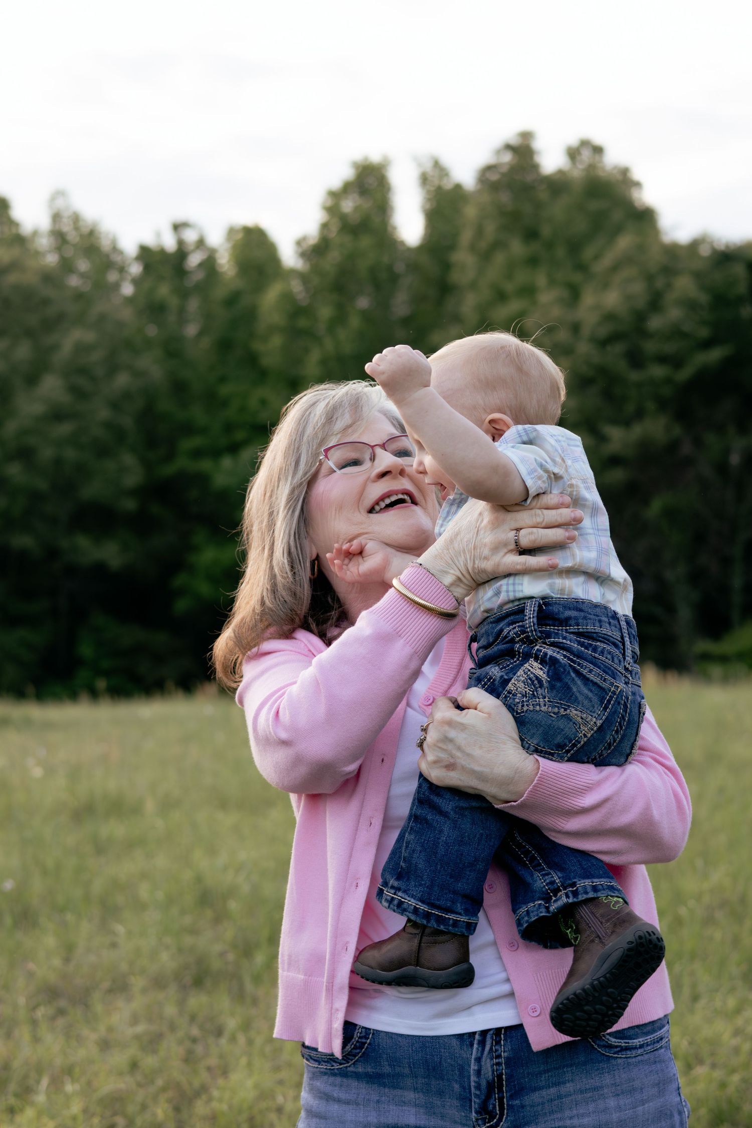 Grandmother laughing joyfully while lifting her baby grandchild outdoors in a green meadow, wearing a pink cardigan.