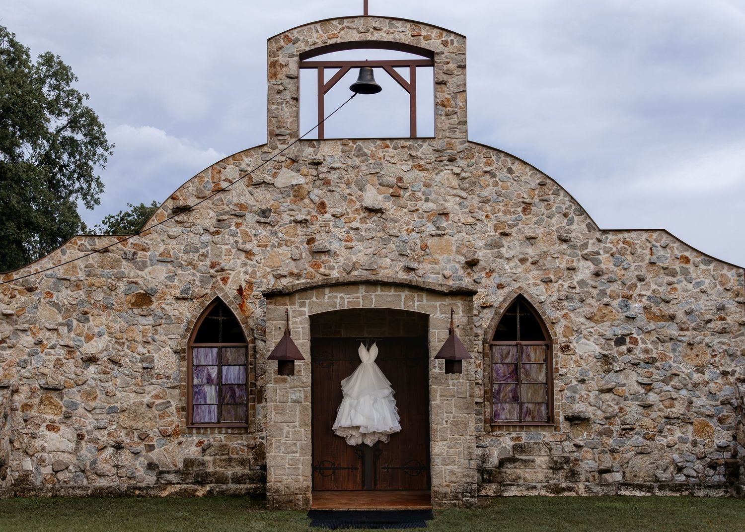 A white wedding dress hangs on a rustic stone chapel entrance with arched windows and a bell tower.