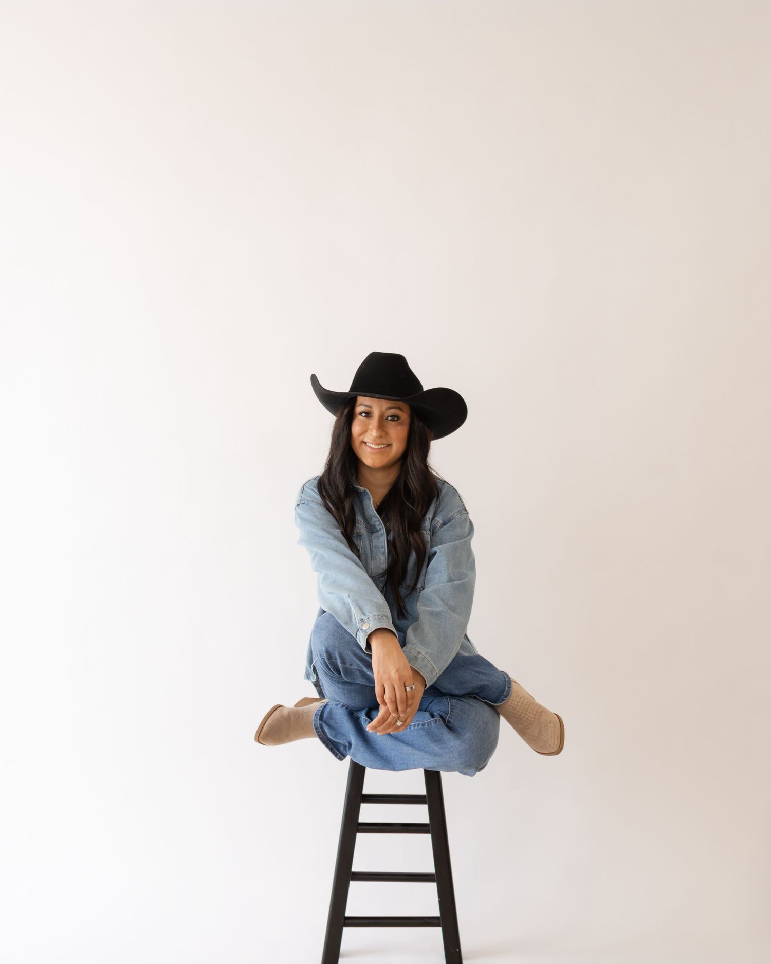 Woman in black cowboy hat, denim shirt, and jeans sits cross-legged on a dark stool against a white background.