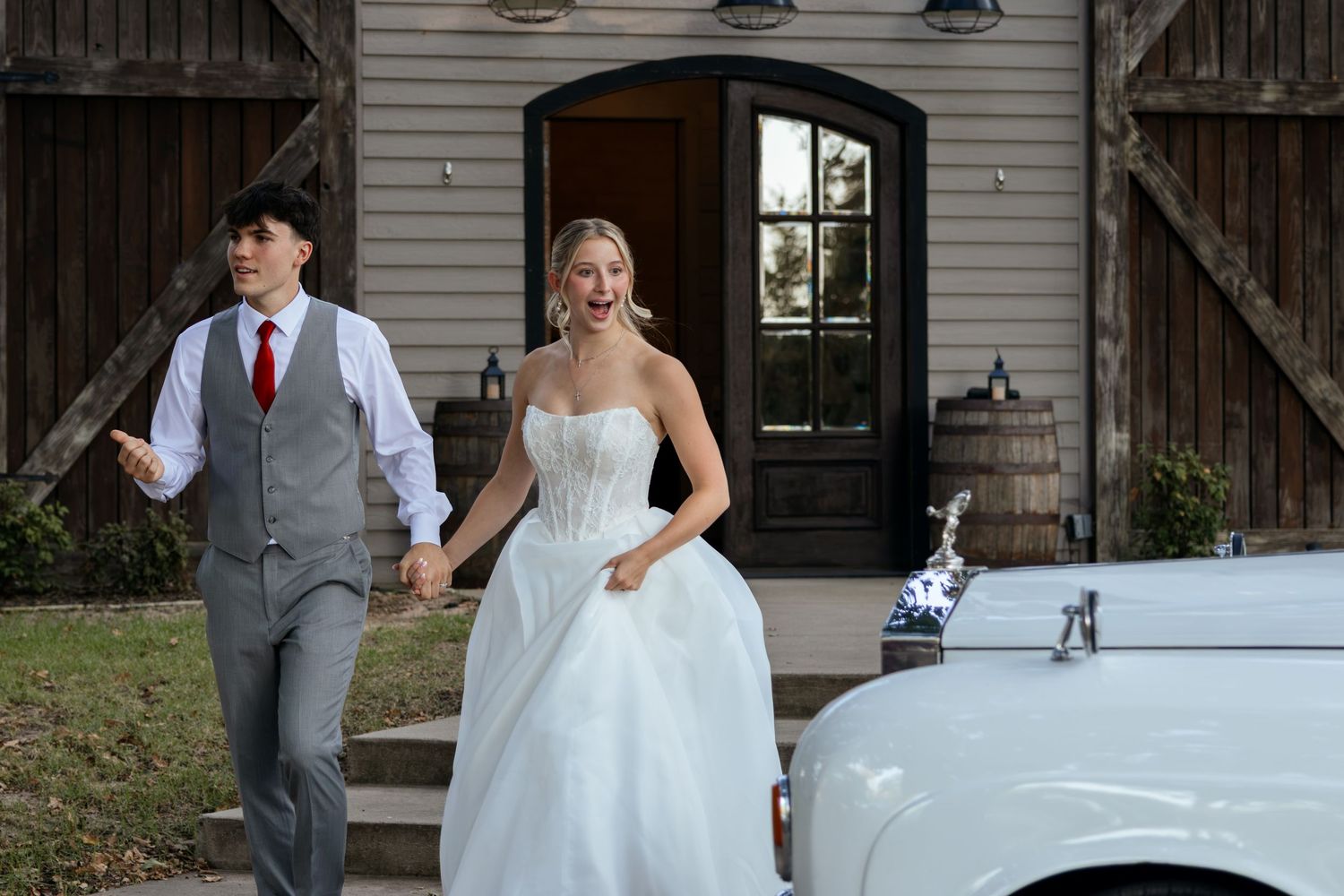 Bride in white strapless gown and groom in gray suit walk hand-in-hand near a vintage white car at a barn venue.