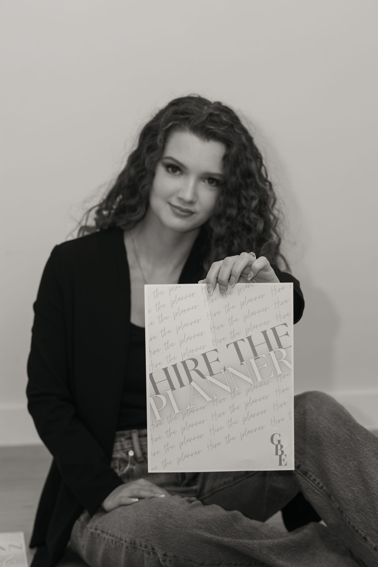 Young woman with curly hair holding a Hire The Planner sign, black and white portrait photo.