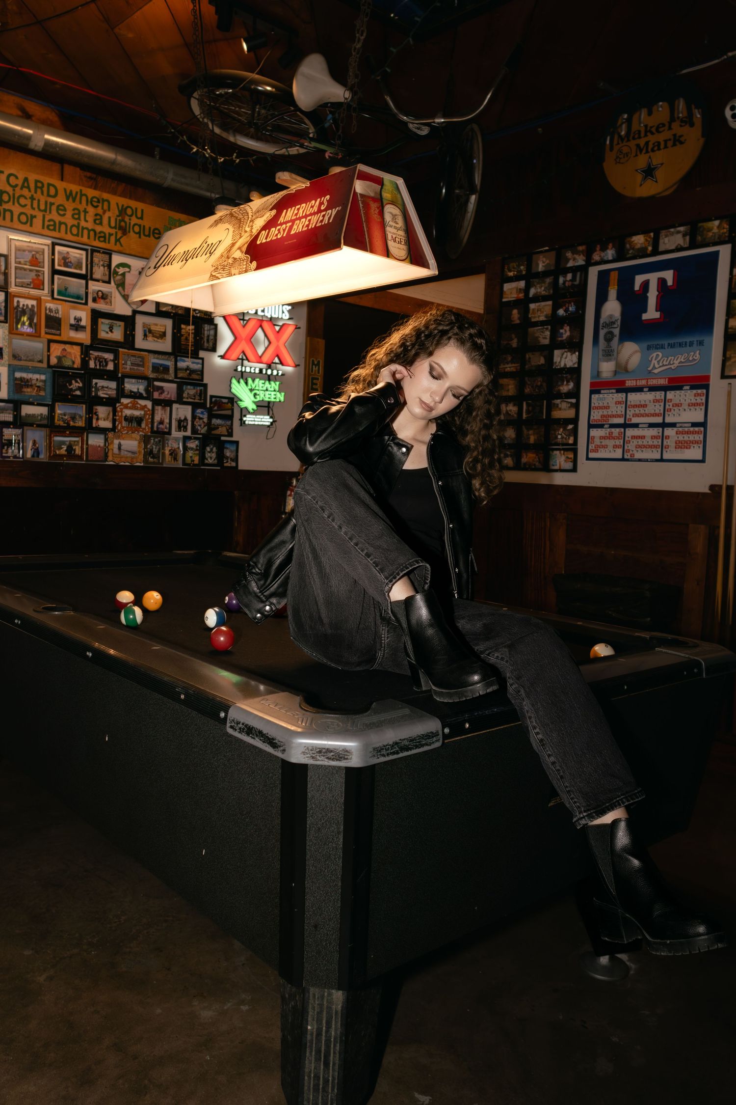Woman in black outfit sitting on a pool table in a dimly lit bar with neon signs and posters.