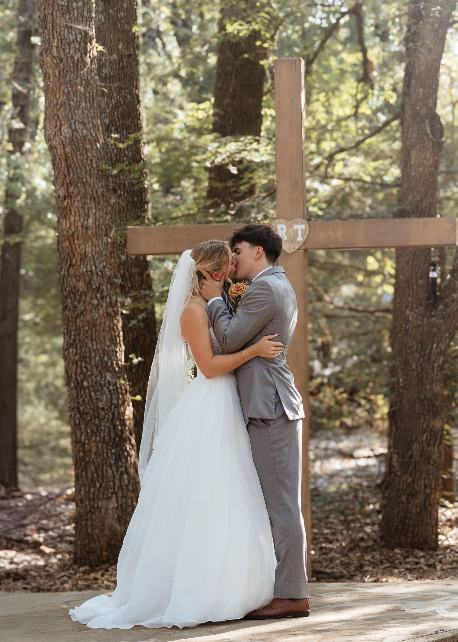 A bride and groom share a first kiss in front of a wooden cross surrounded by tall pine trees.