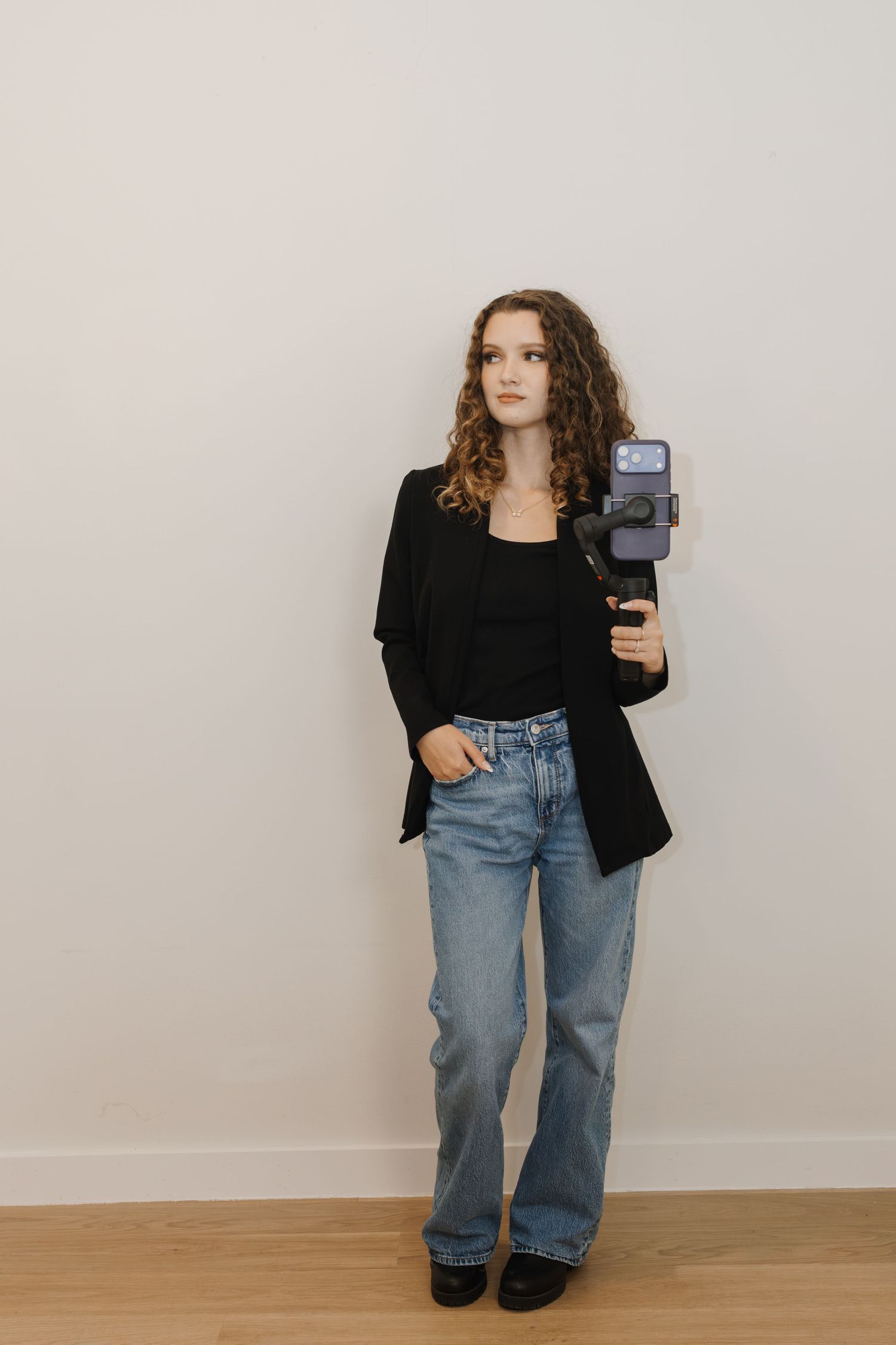 Young woman with curly hair in black blazer and jeans holding a phone gimbal stabilizer.