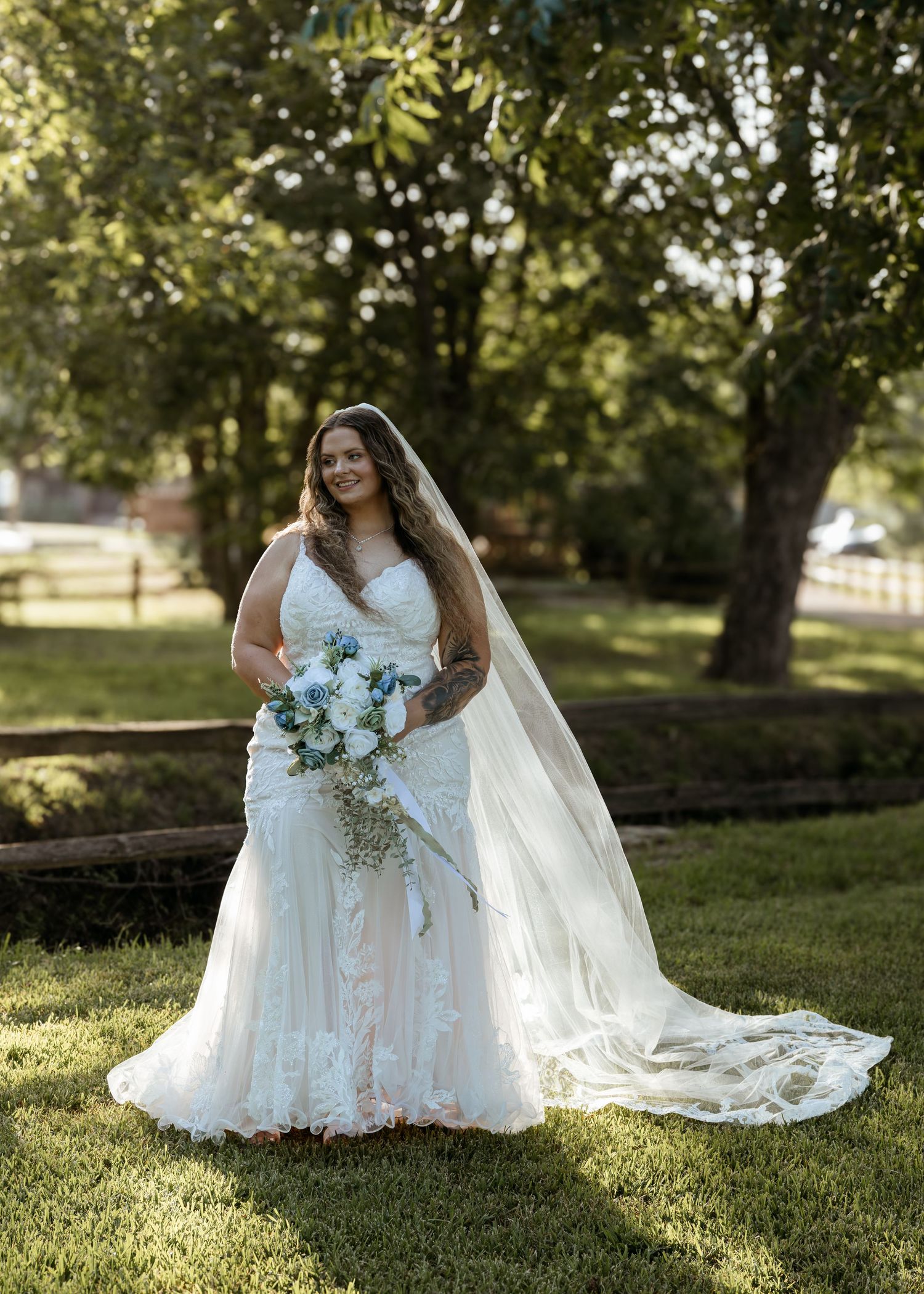 Smiling bride in lace mermaid gown with veil holding bouquet outdoors on green lawn.