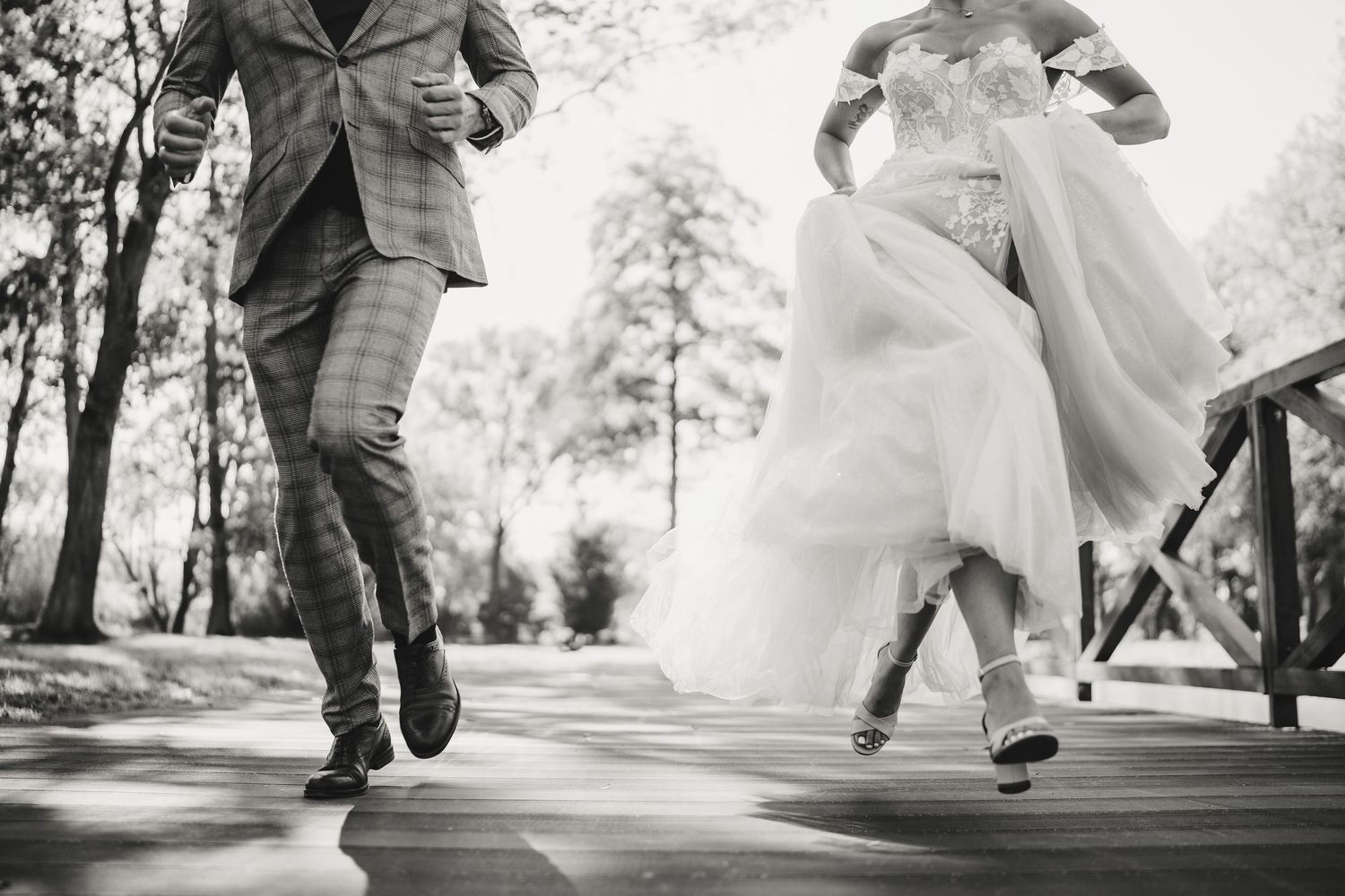 Black and white photo of playful newlyweds dancing on a wooden bridge.
