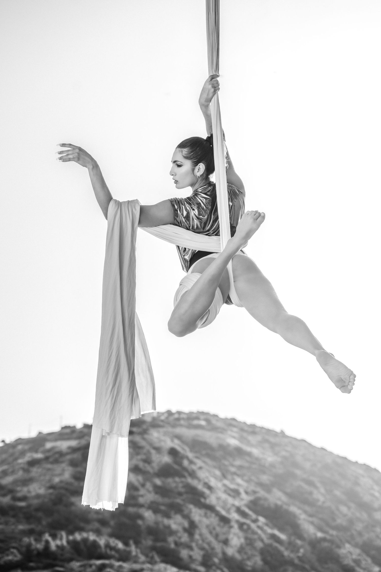 Black and white photo of aerial silk dancer suspended mid-air outdoors, performing graceful split pose against mountain backdrop.