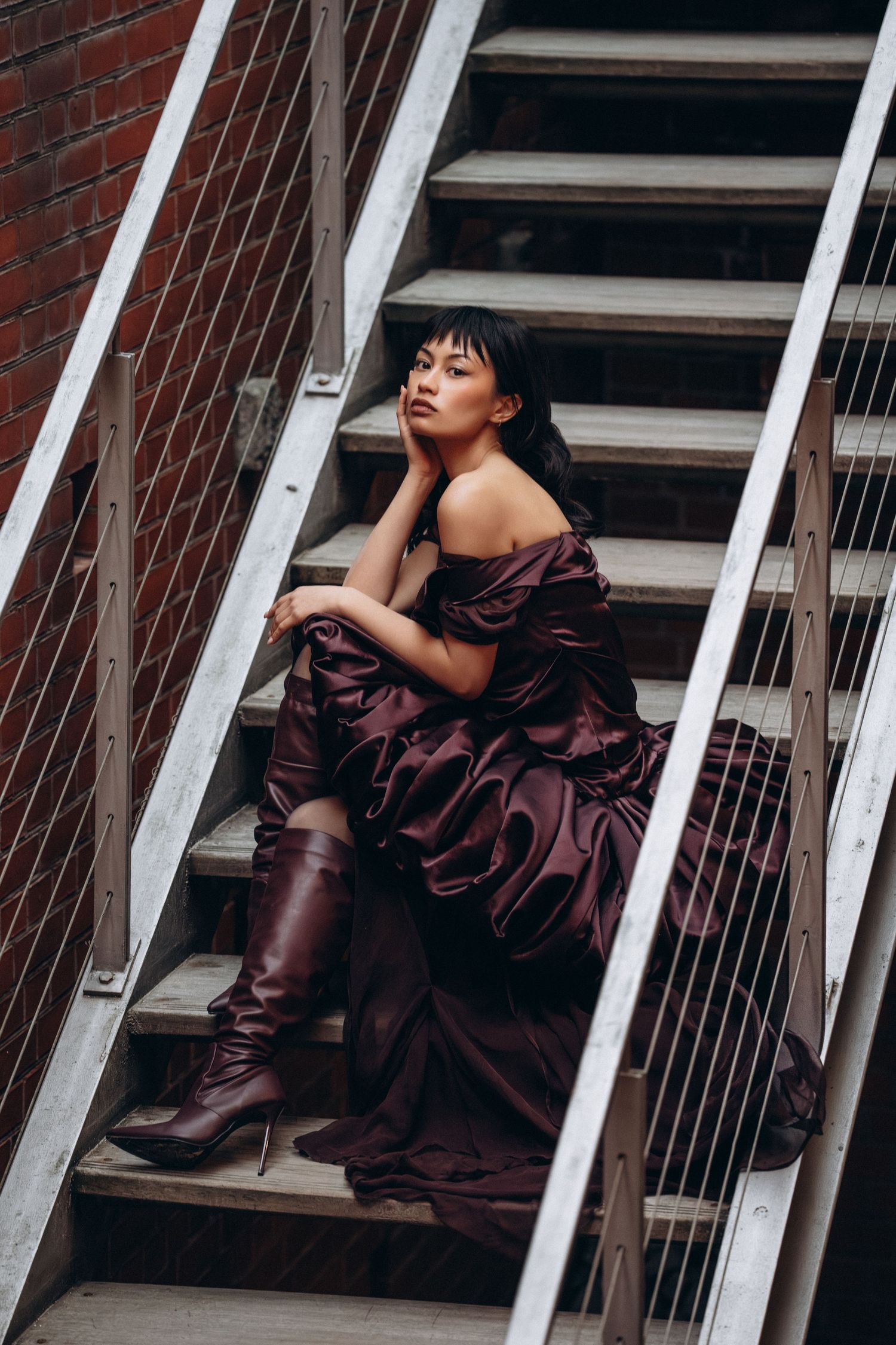 A person in a burgundy gown and tall boots poses dramatically on industrial metal stairs against a brick wall.