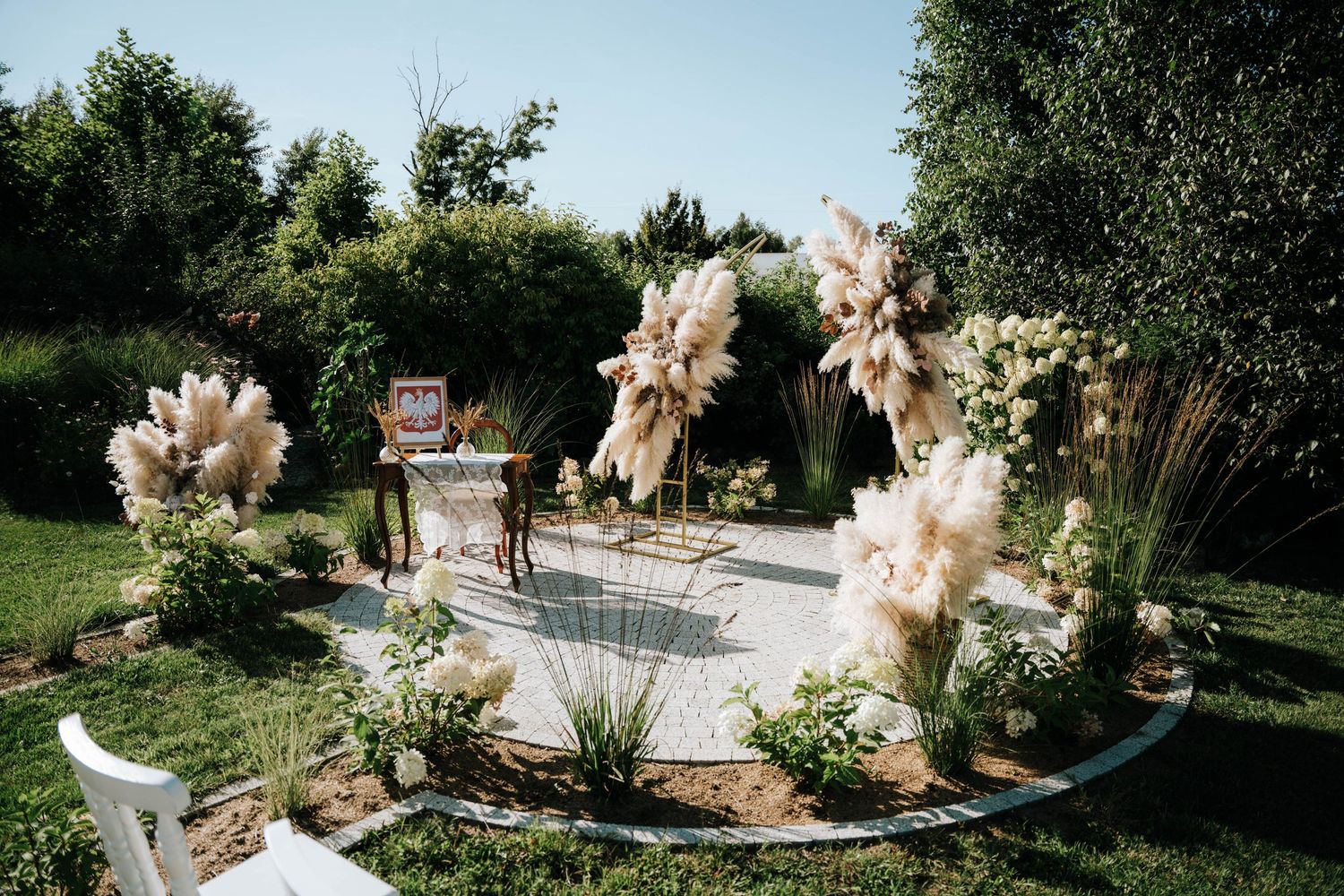 Outdoor wedding ceremony setup with pampas grass arrangements, white floral decor, and lush greenery in a garden setting.