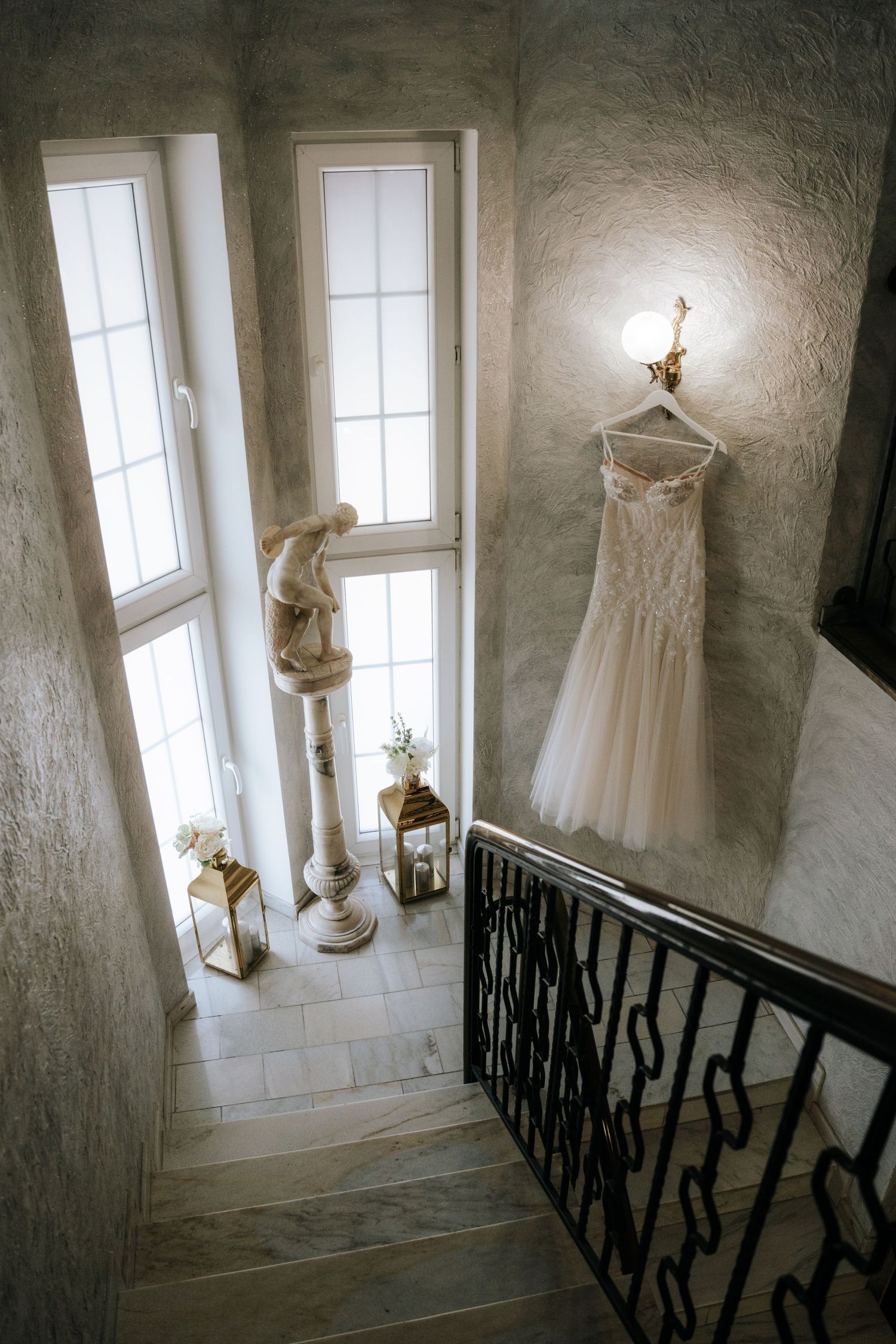 Elegant wedding dress hanging on staircase railing in a grand foyer with marble statue and ornate wall sconce lighting.