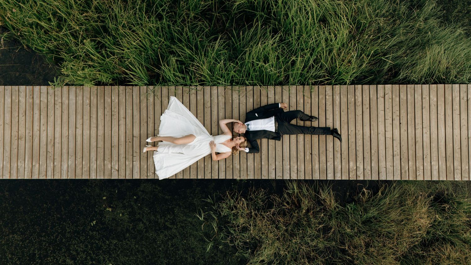 Aerial view of bride and groom lying on wooden boardwalk surrounded by lush green grass on wedding day.
