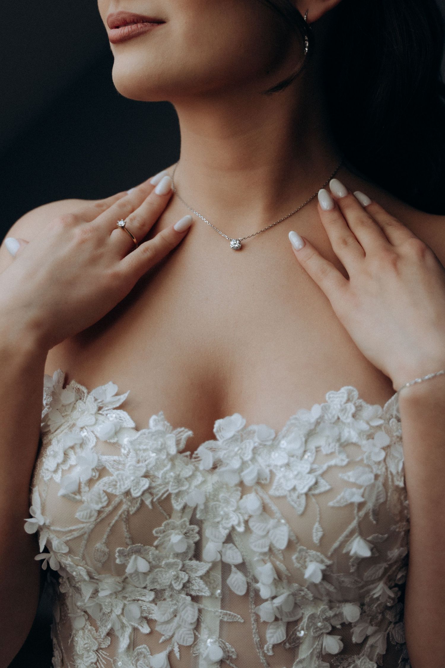 Bride in floral lace wedding gown touching delicate diamond necklace, wearing ring, manicured nails, elegant bridal jewelry.