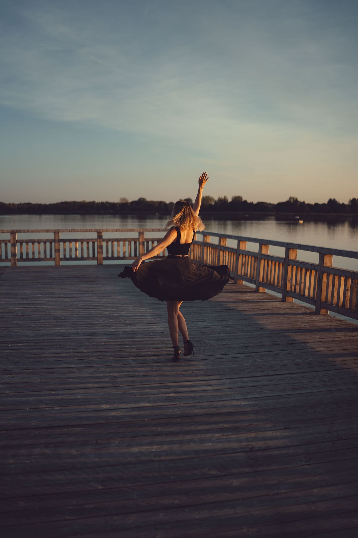 Dancer in flowing dress poses gracefully on lakeside boardwalk at sunset.
