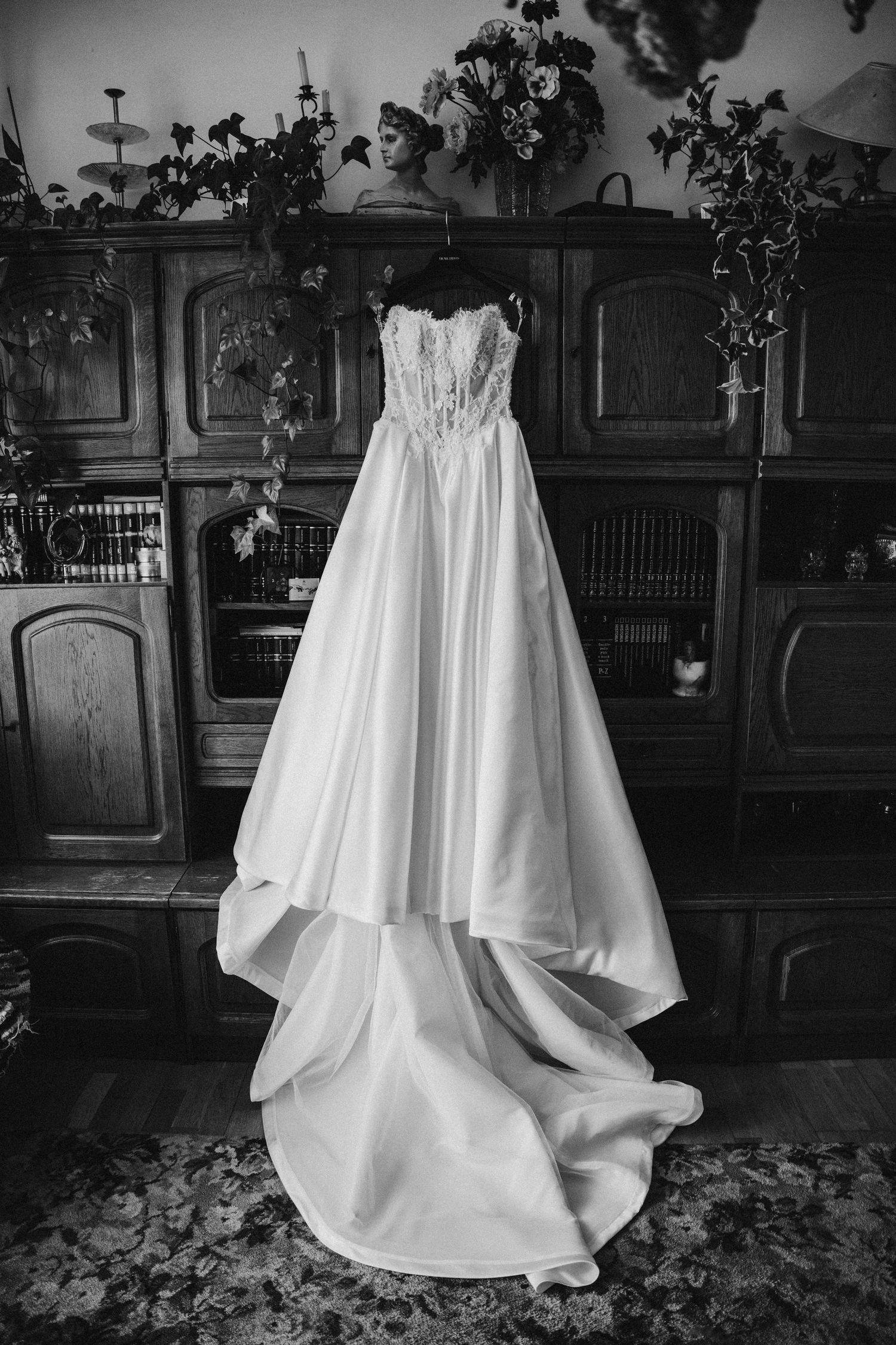 Black and white photo of an elegant strapless wedding gown hanging on a wooden cabinet with floral arrangements.