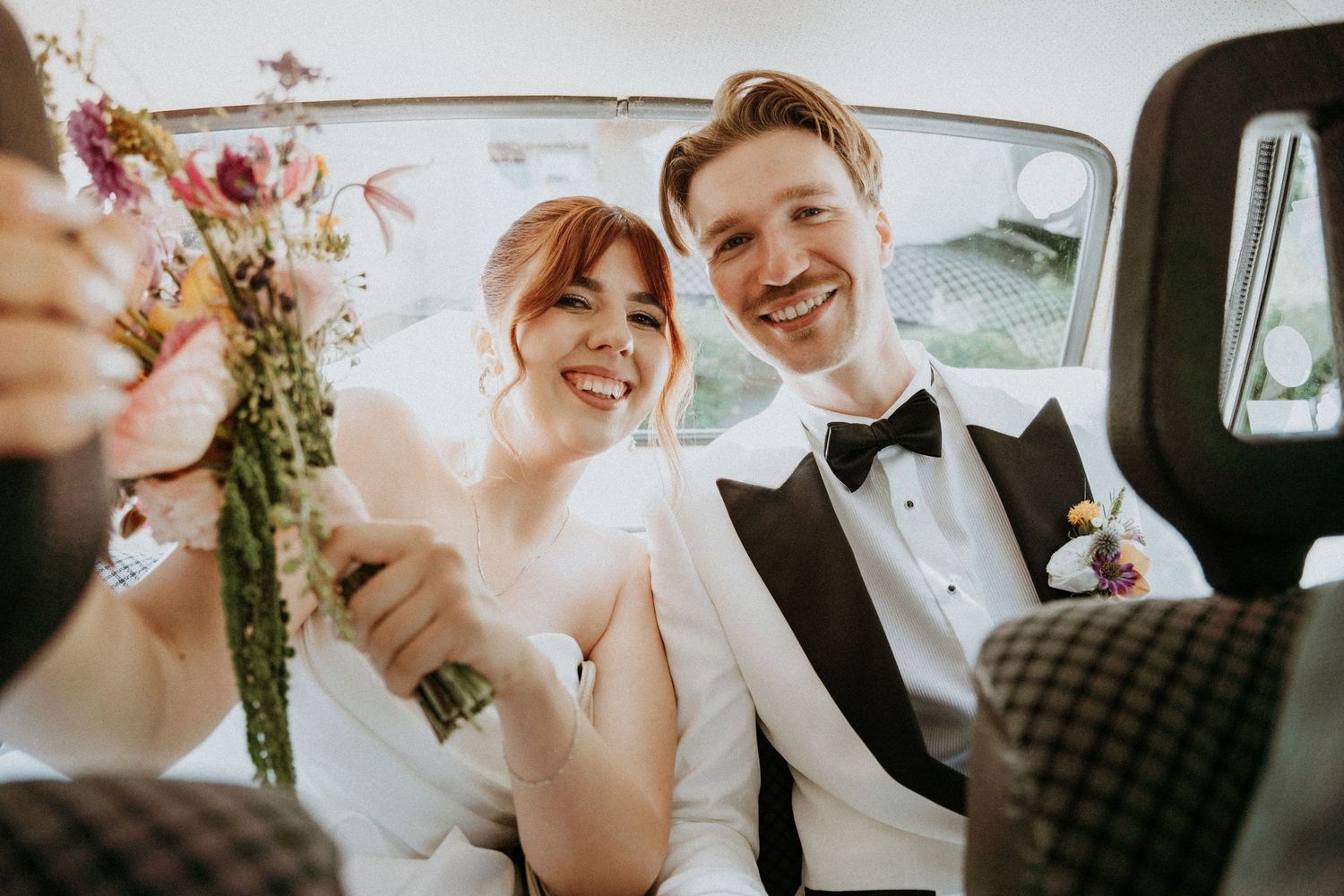 Happy newlywed couple sitting in a vintage car, bride holding a floral bouquet, groom in a black tuxedo.