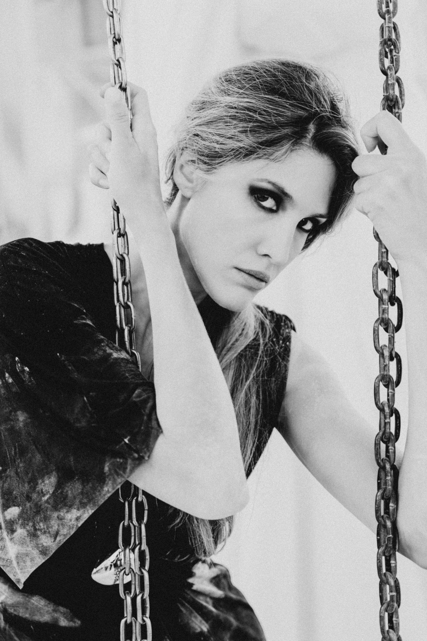 Black and white portrait of a blonde woman with dramatic eye makeup sitting on a chain swing, gazing intensely at camera.