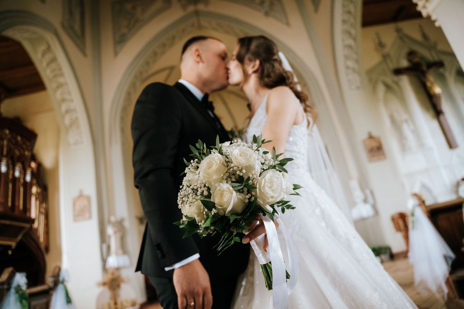 Bride and groom share a romantic kiss in an elegant church, bride holding a white rose bouquet.