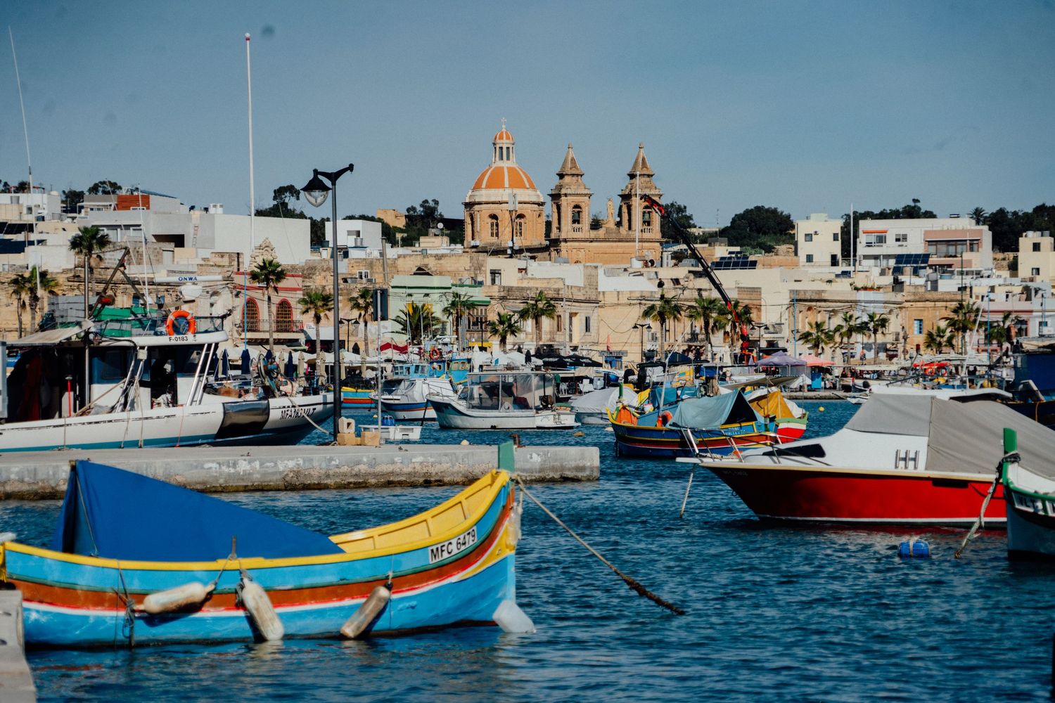 Vibrant Maltese fishing boats called Luzzus float in a picturesque harbor with church domes visible.