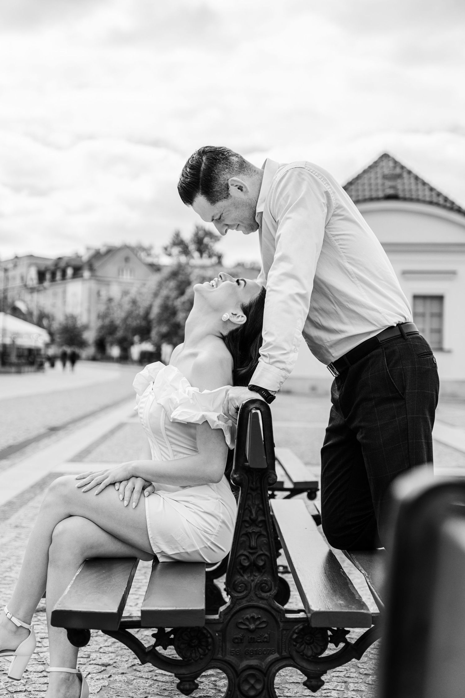 A romantic black and white photo of a couple sharing an intimate moment while sitting on an ornate bench in a city square.