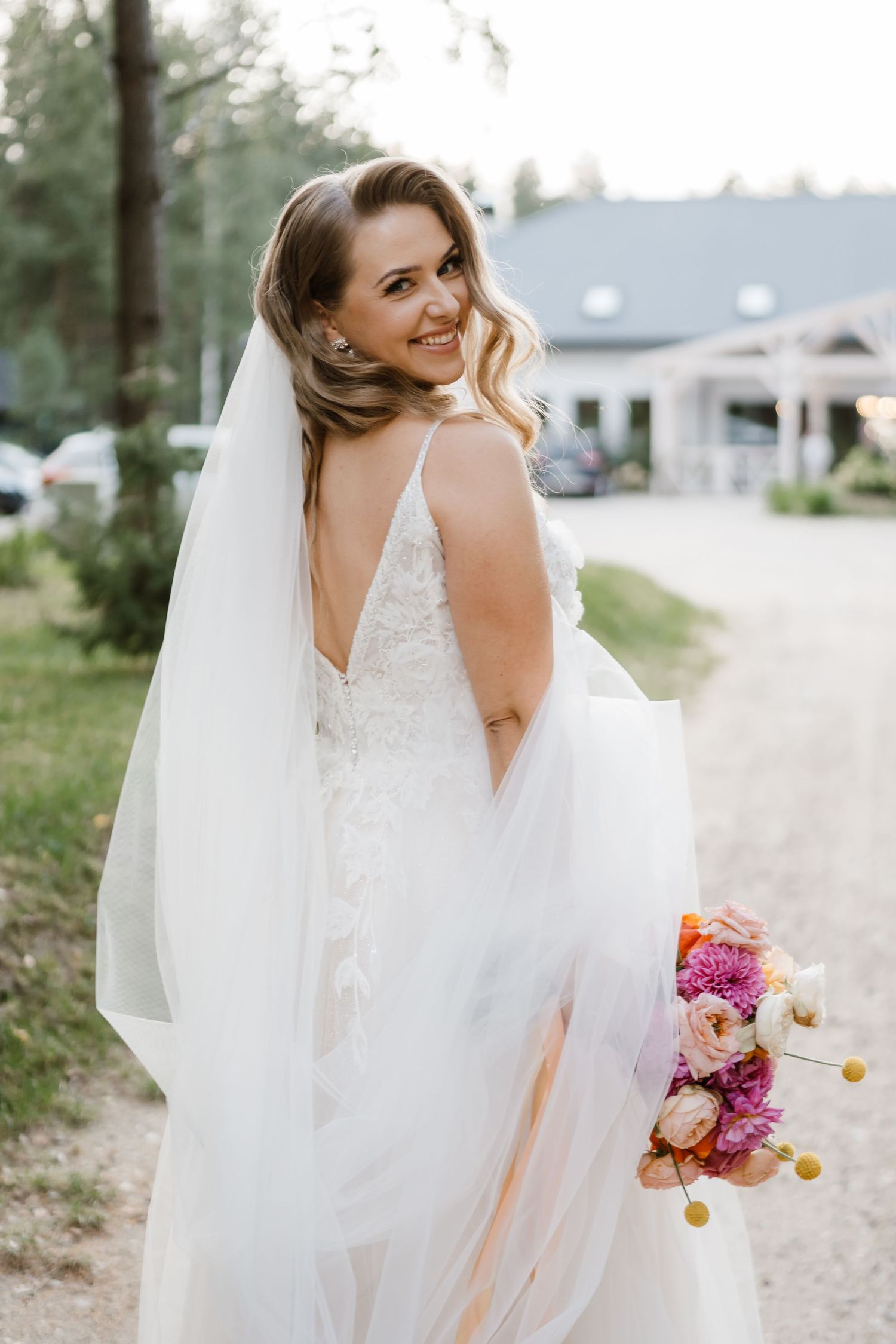 A bride wearing a lace wedding dress holds a colorful bouquet while posing outdoors.