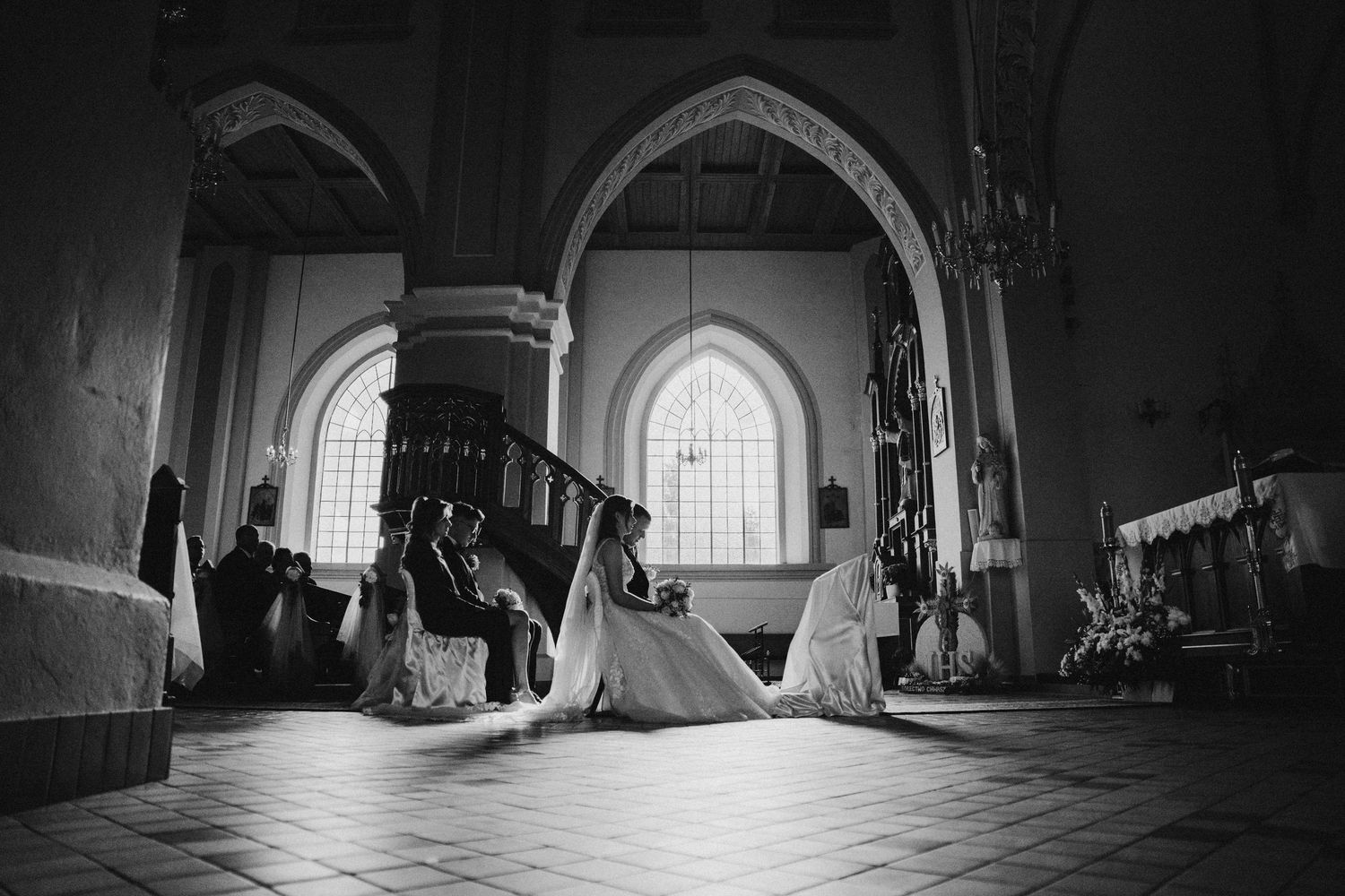 Black and white photo of a bride kneeling in prayer inside a grand church with arched windows and ornate wooden pews.