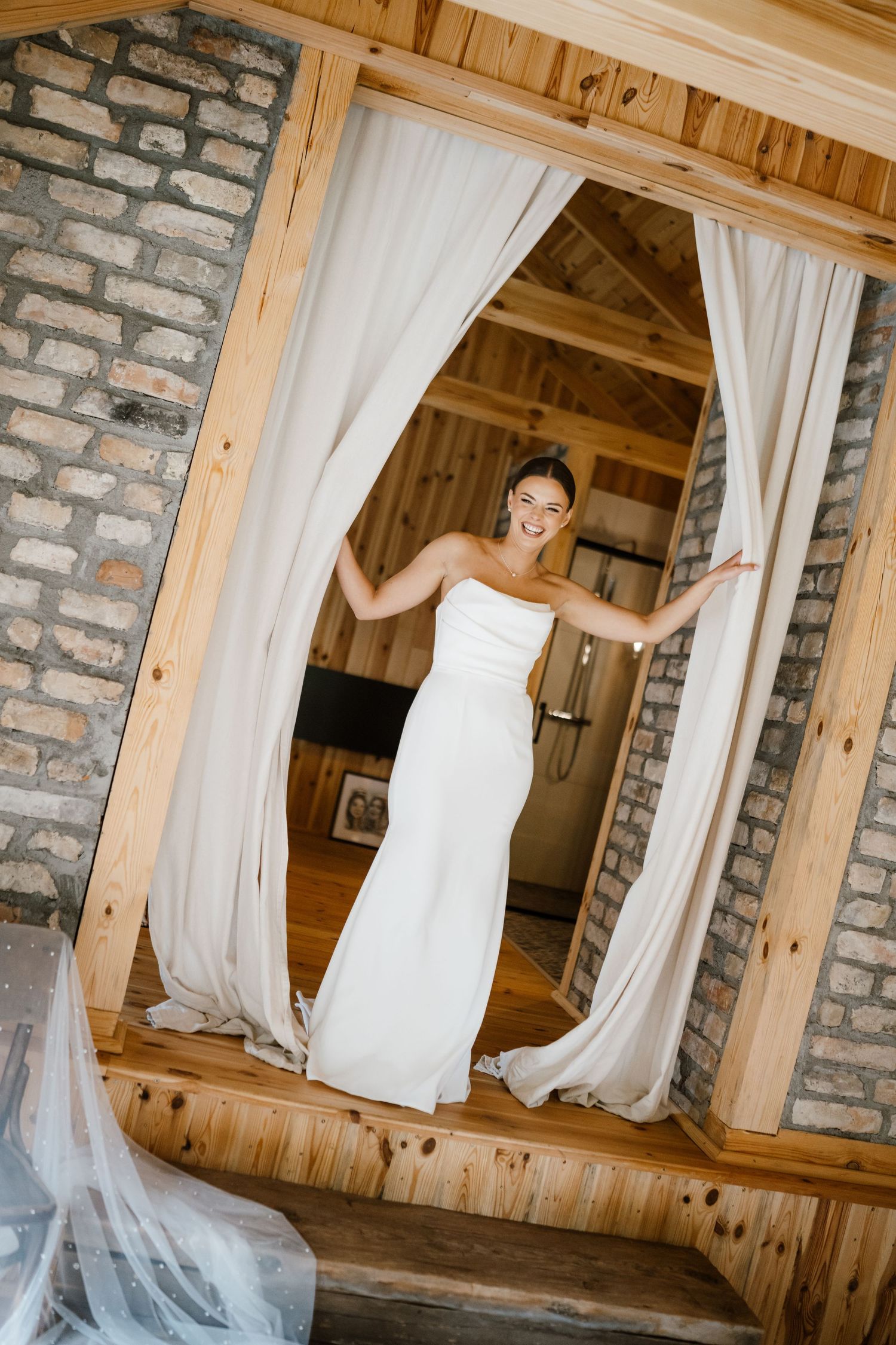 A wedding dress displayed in a rustic venue with exposed brick walls and flowing white curtains.