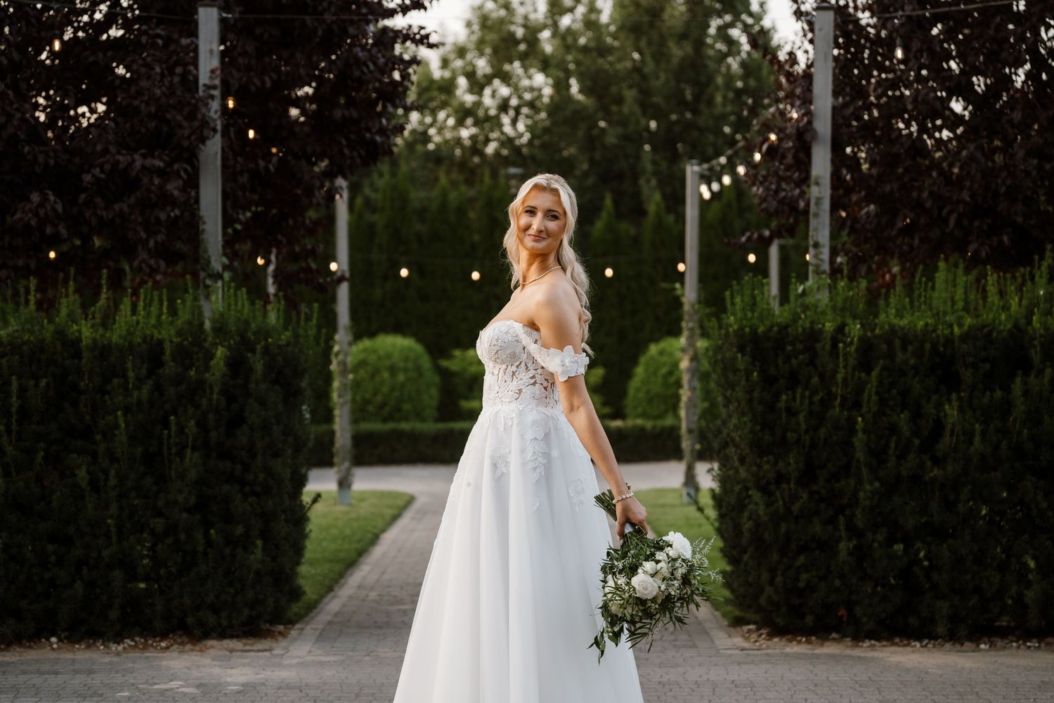 A bride in a flowing white gown poses elegantly in a garden pathway lined with manicured hedges.