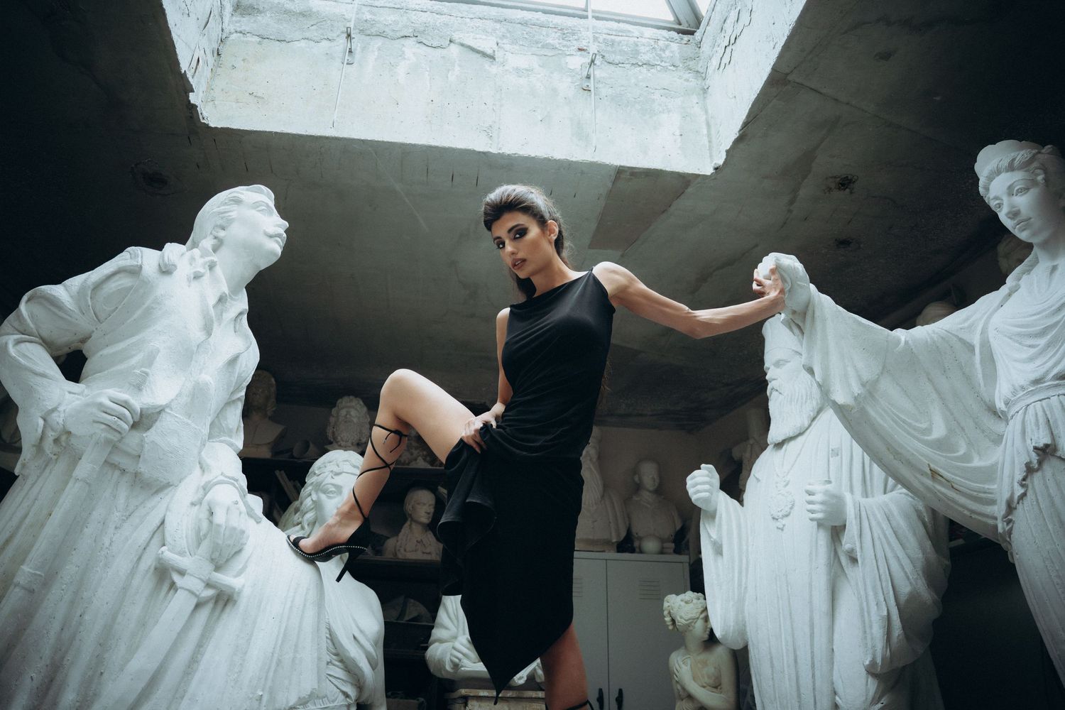Woman in black dress poses among white classical sculptures in a dramatic studio setting.