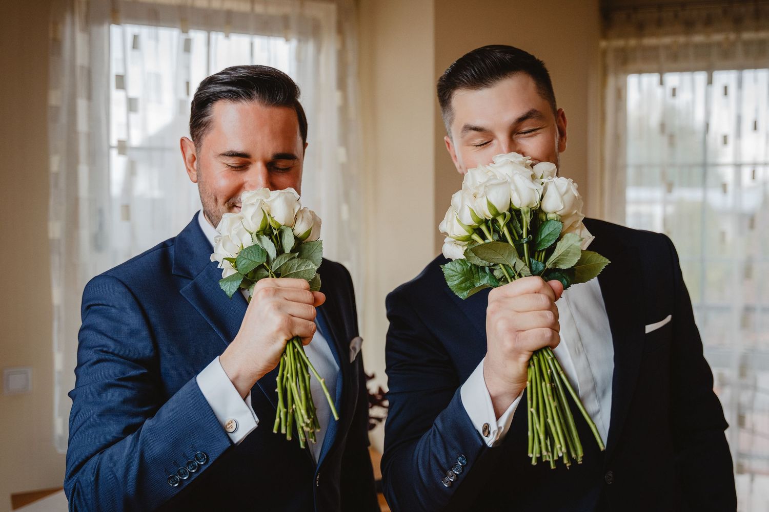 Groomsmen in navy suits smell their bouquets of white flowers while standing in a row during a wedding ceremony.
