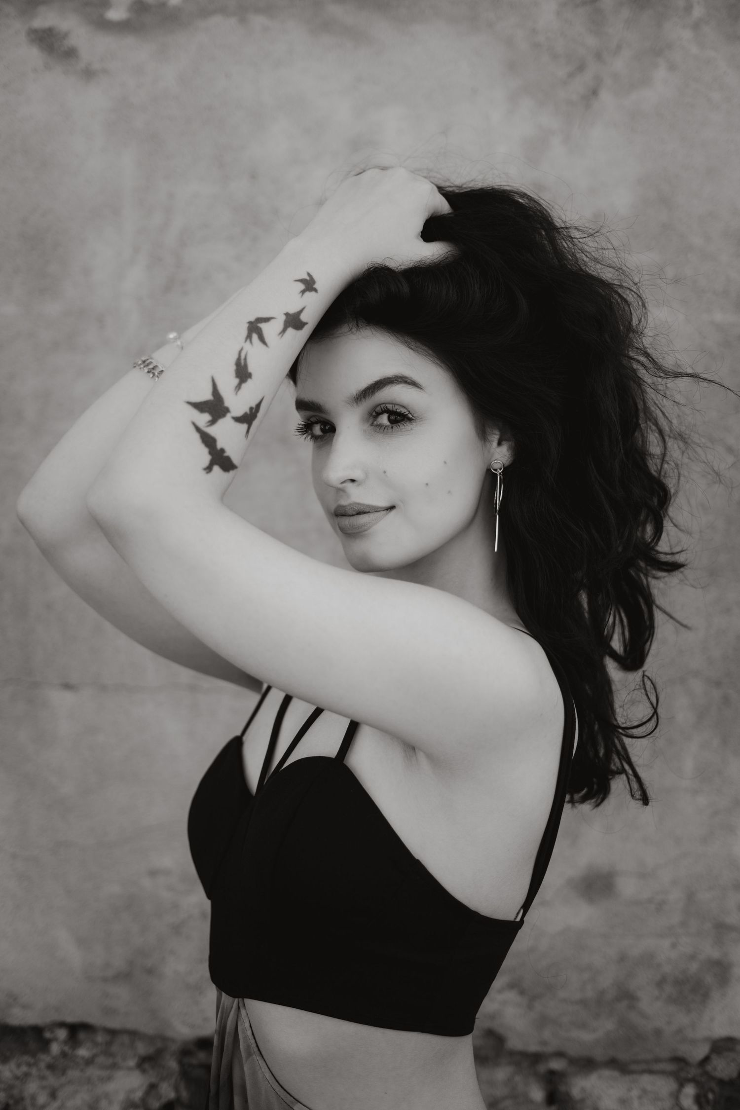 Black and white portrait of a person with dark flowing hair wearing a black top and showing a bird tattoo on their arm.