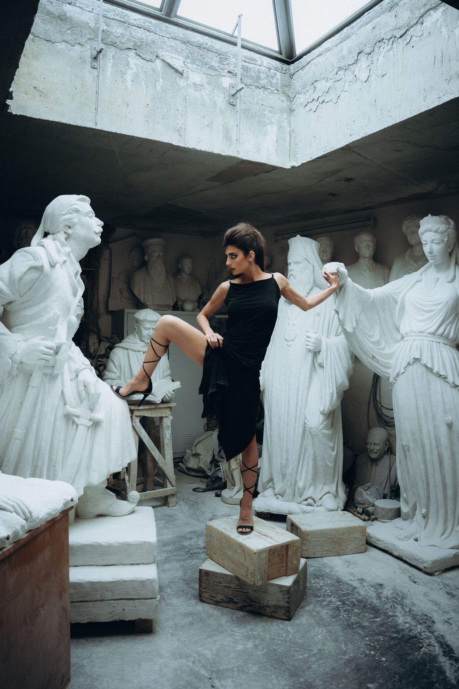 Woman in black dress poses elegantly among white classical sculptures in a dimly lit studio.