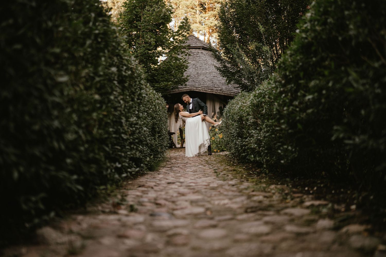 A romantic garden path lined with lush hedges leads to a rustic thatched-roof cottage in soft evening light.
