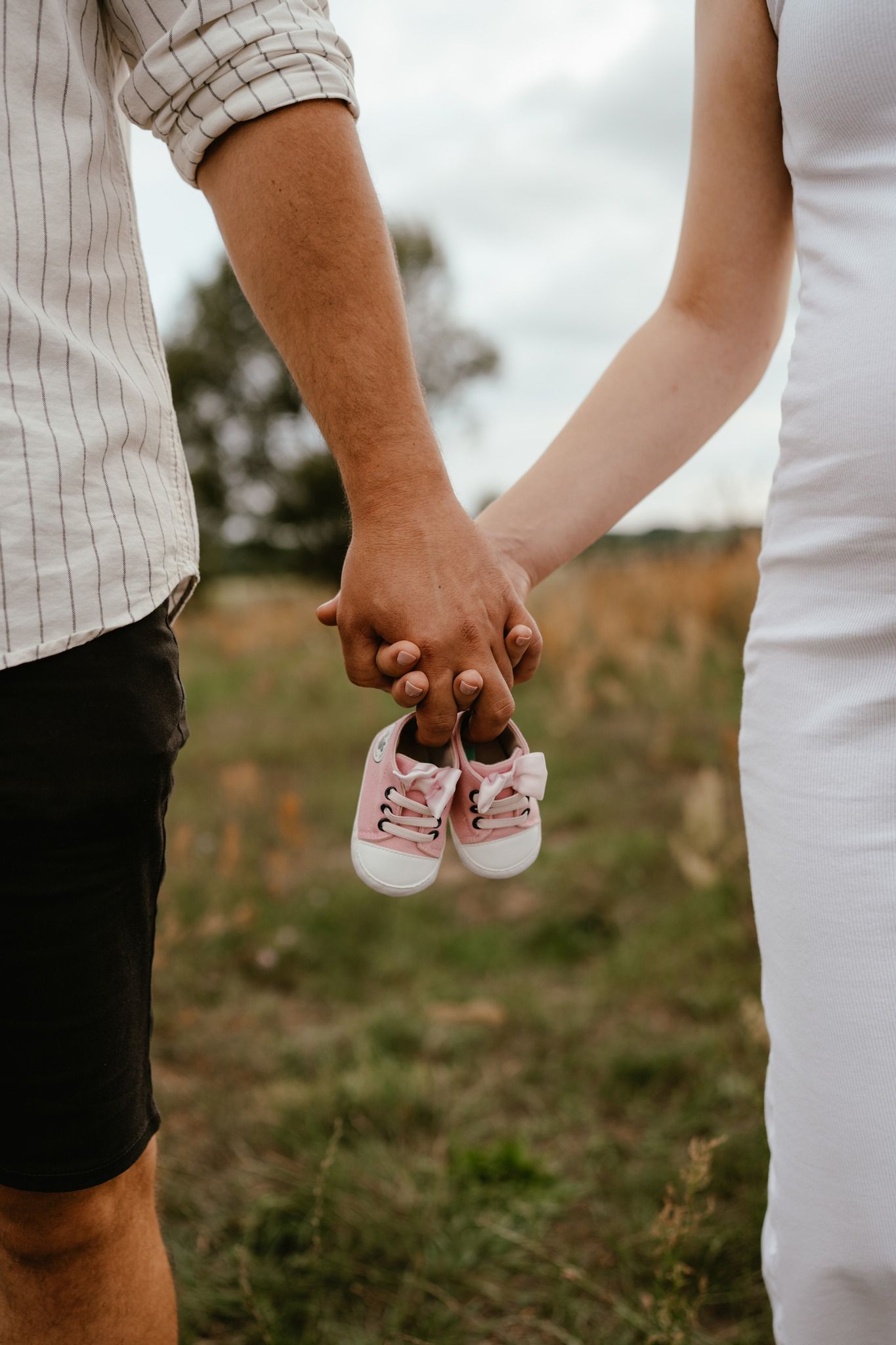 Couple holding hands and tiny pink baby shoes in a field, announcing pregnancy outdoors.