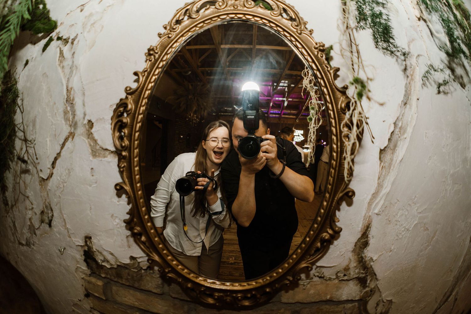 A series of vintage gold ornate mirrors mounted on a distressed white wall reflect figures taking selfie photos.