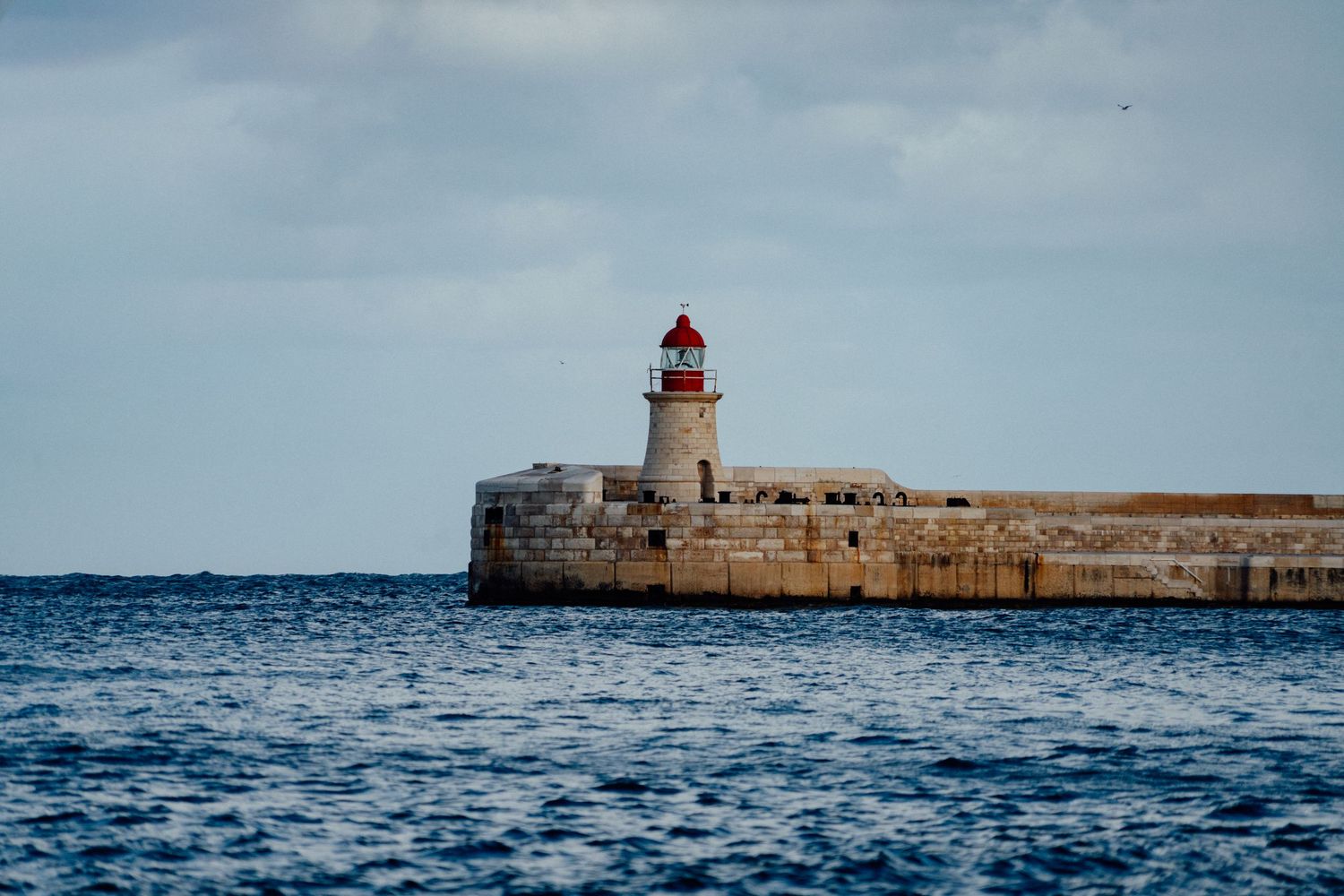 Historic stone lighthouse with red top stands at the end of a breakwater in the Mediterranean Sea.