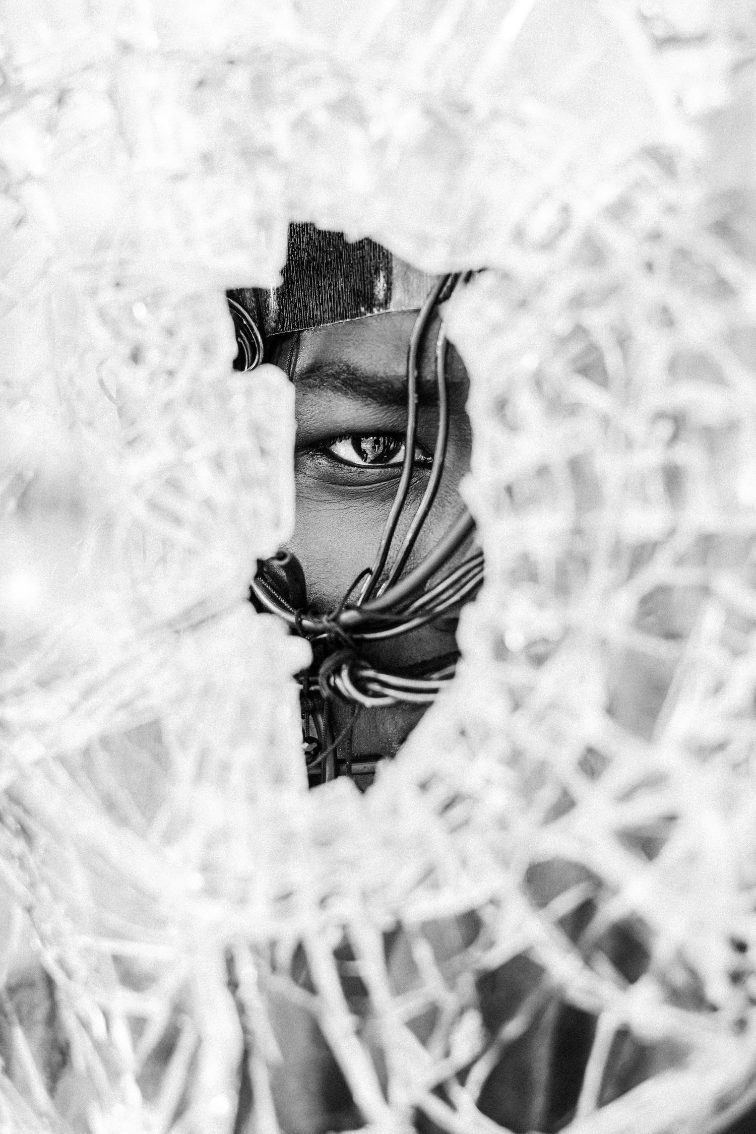 Black and white photo of a person peering through a hole in shattered broken glass, one eye visible.