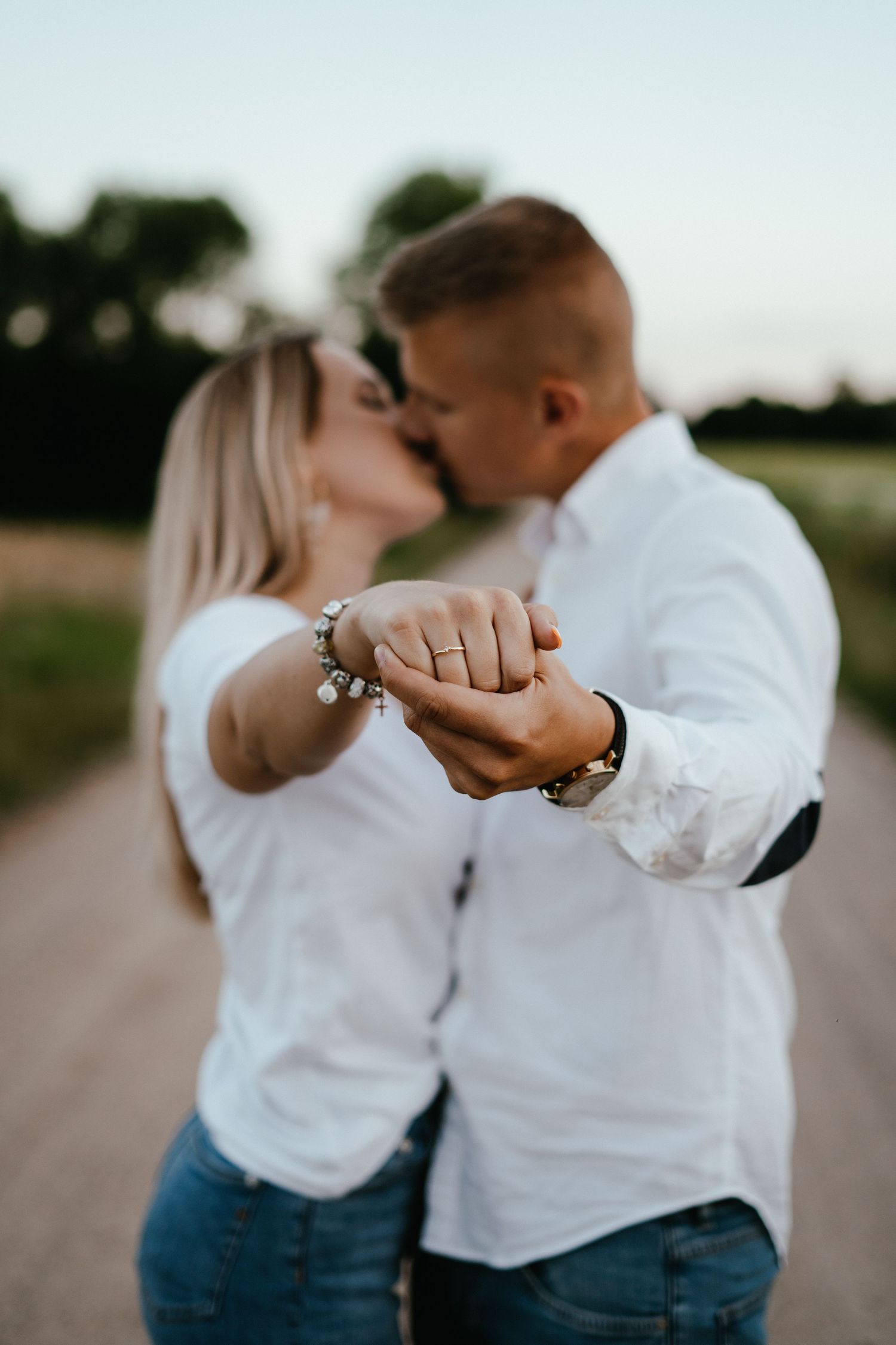A couple in white shirts and jeans hold hands and embrace while showing off an engagement ring in a romantic photoshoot.