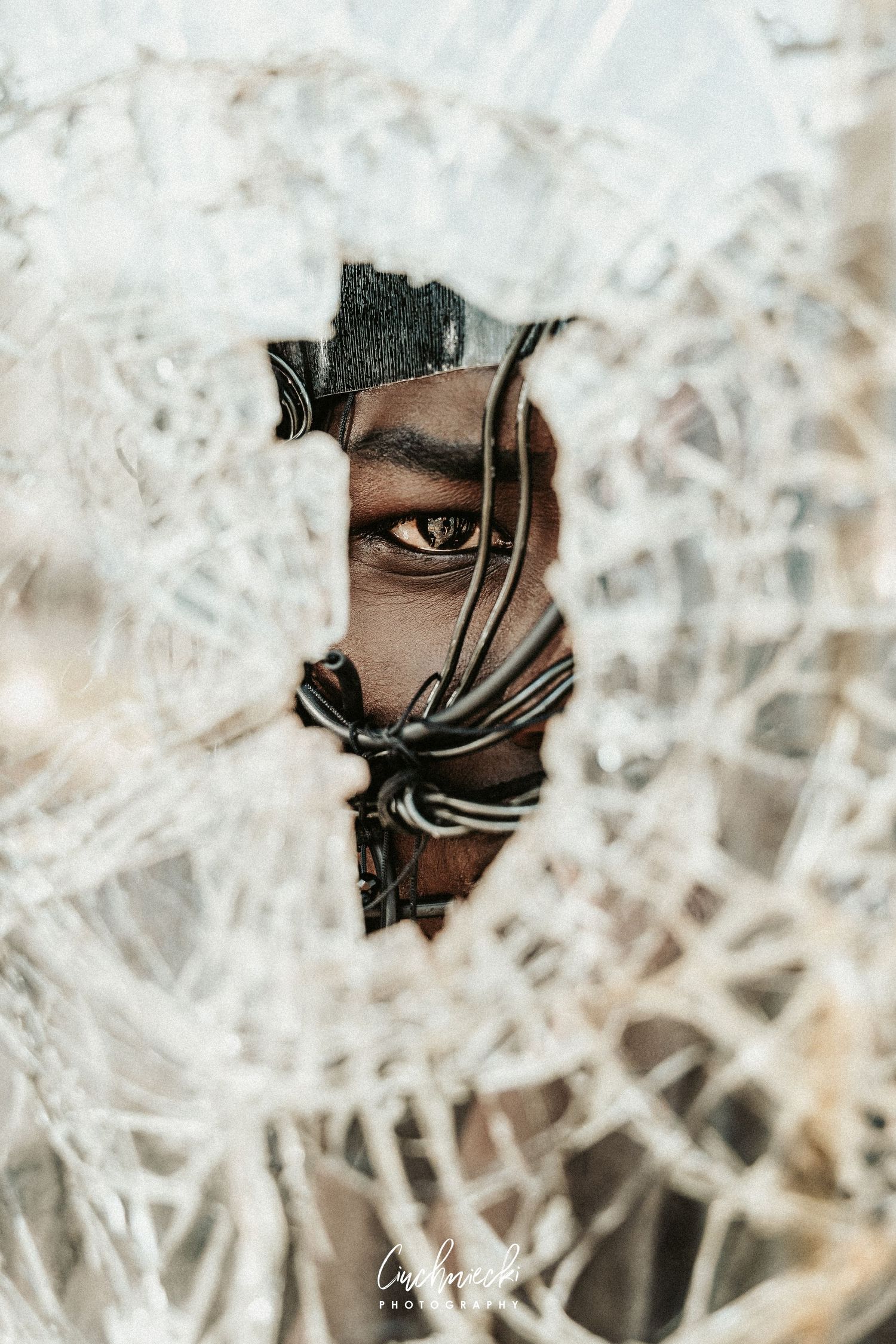 A person peers through a shattered glass hole, with cracked glass surrounding their eye and face in a dramatic close-up.